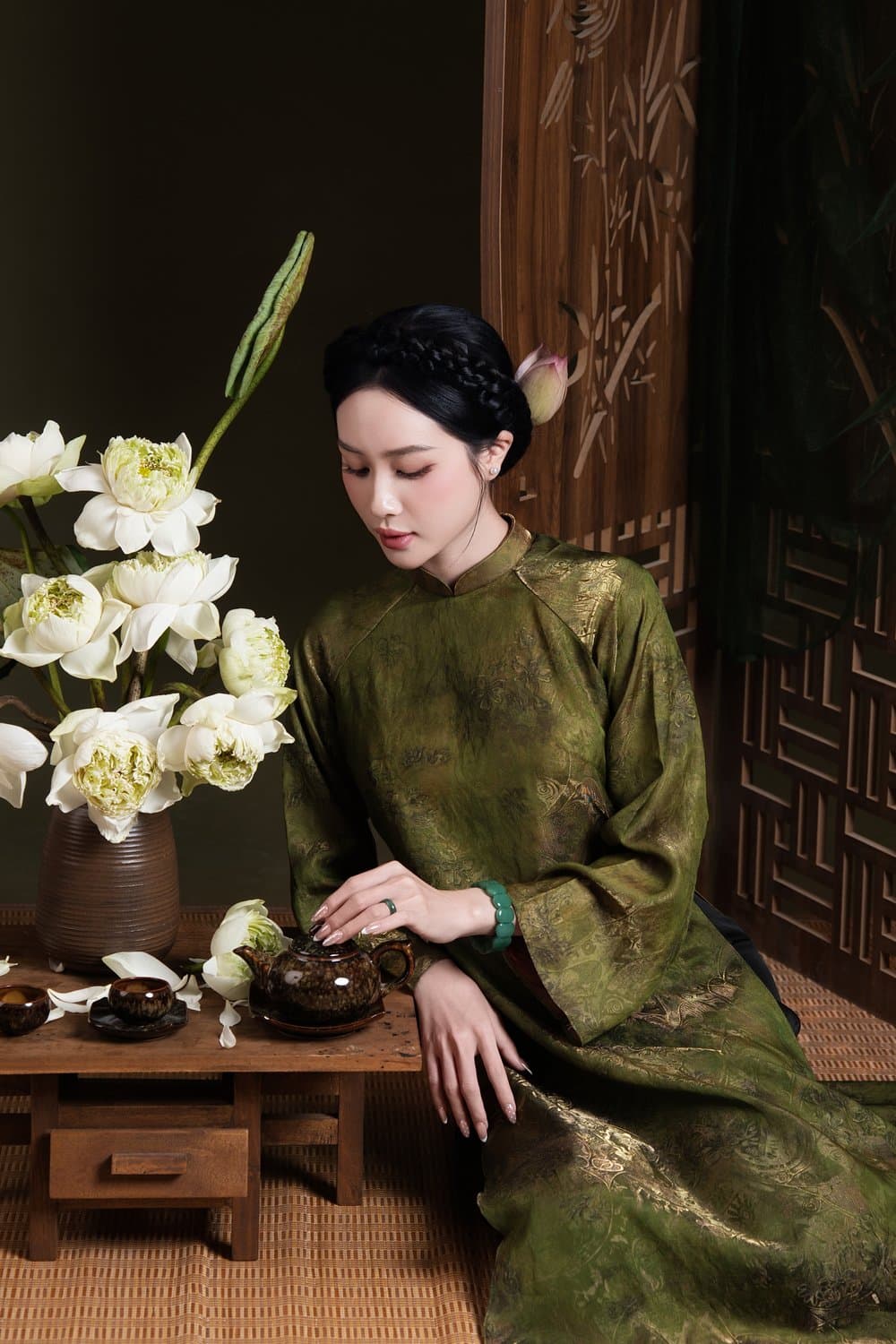 Áo dài xanh rêu ngồi bên bàn gỗ trầm, sen trắng — concept sen studio Xanh rêu cổ điển