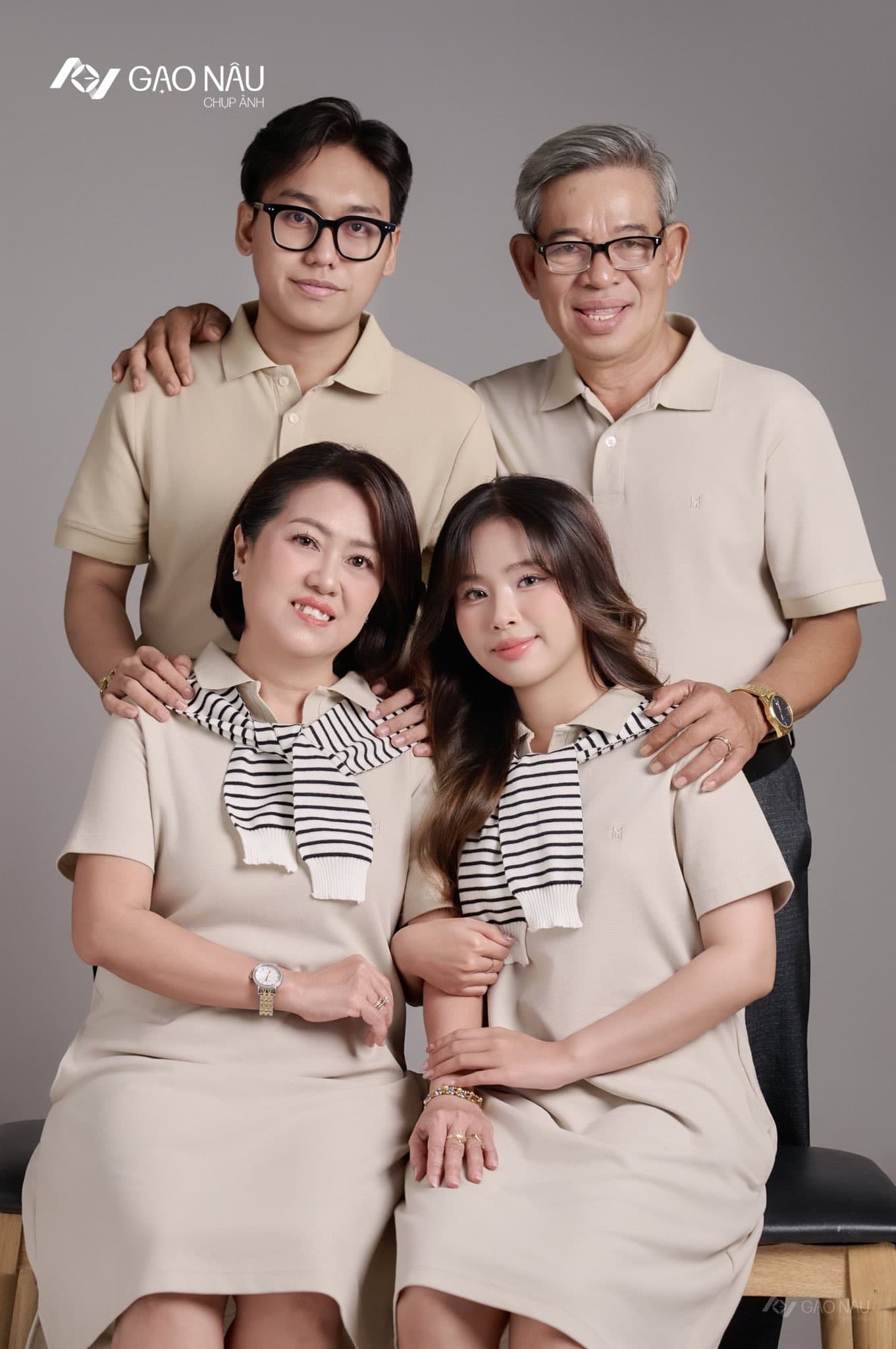 Cận cảnh gia đình bốn người trong tone be — mẹ và con gái ngồi ở ghế giữa, cùng khoác khăn sọc đen trắng giống hệt nhau; ba và em trai đeo kính đứng phía sau, đặt tay lên vai hai người phụ nữ. Tất cả cười ấm.