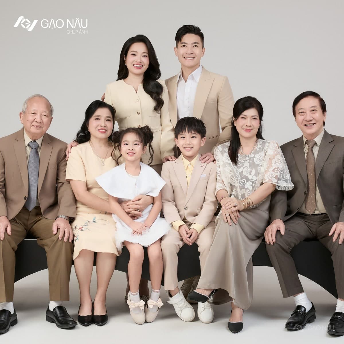 Đại gia đình tám người ba thế hệ đứng quây trong studio — ông bà nội, ông bà ngoại, vợ chồng trẻ đứng sau, hai cháu nhỏ đứng giữa, tone trang phục be nâu