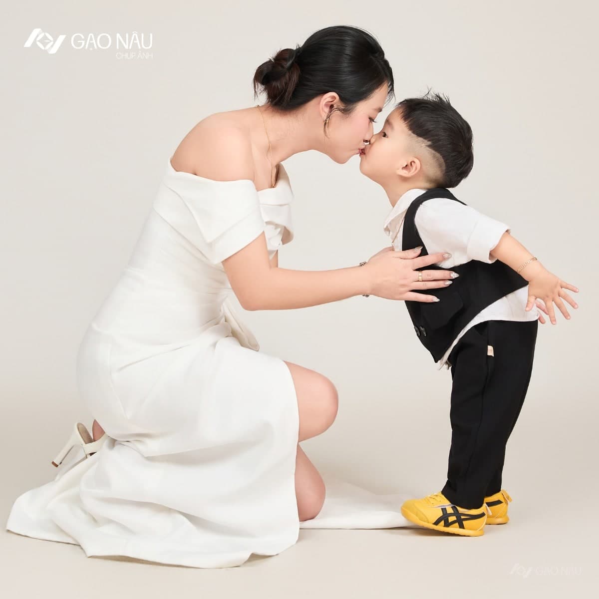 Mẹ mặc đầm trắng quỳ xuống, thơm lên trán con trai hai tuổi mặc áo sơ mi trắng và vest đen, con đi đôi giày thể thao vàng