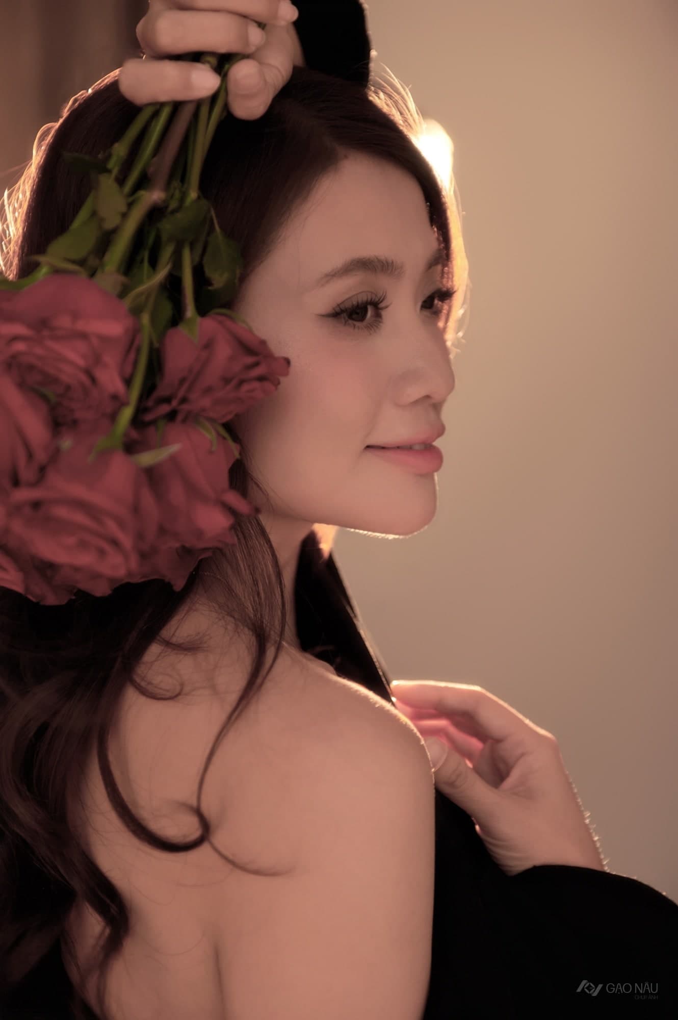 Cô đưa bó hoa hồng đỏ lên ngang mặt, bàn tay mảnh để trên cánh hoa. Ánh sáng vàng ấm phủ toàn khung hình. Một khoảnh khắc dịu và riêng tư.