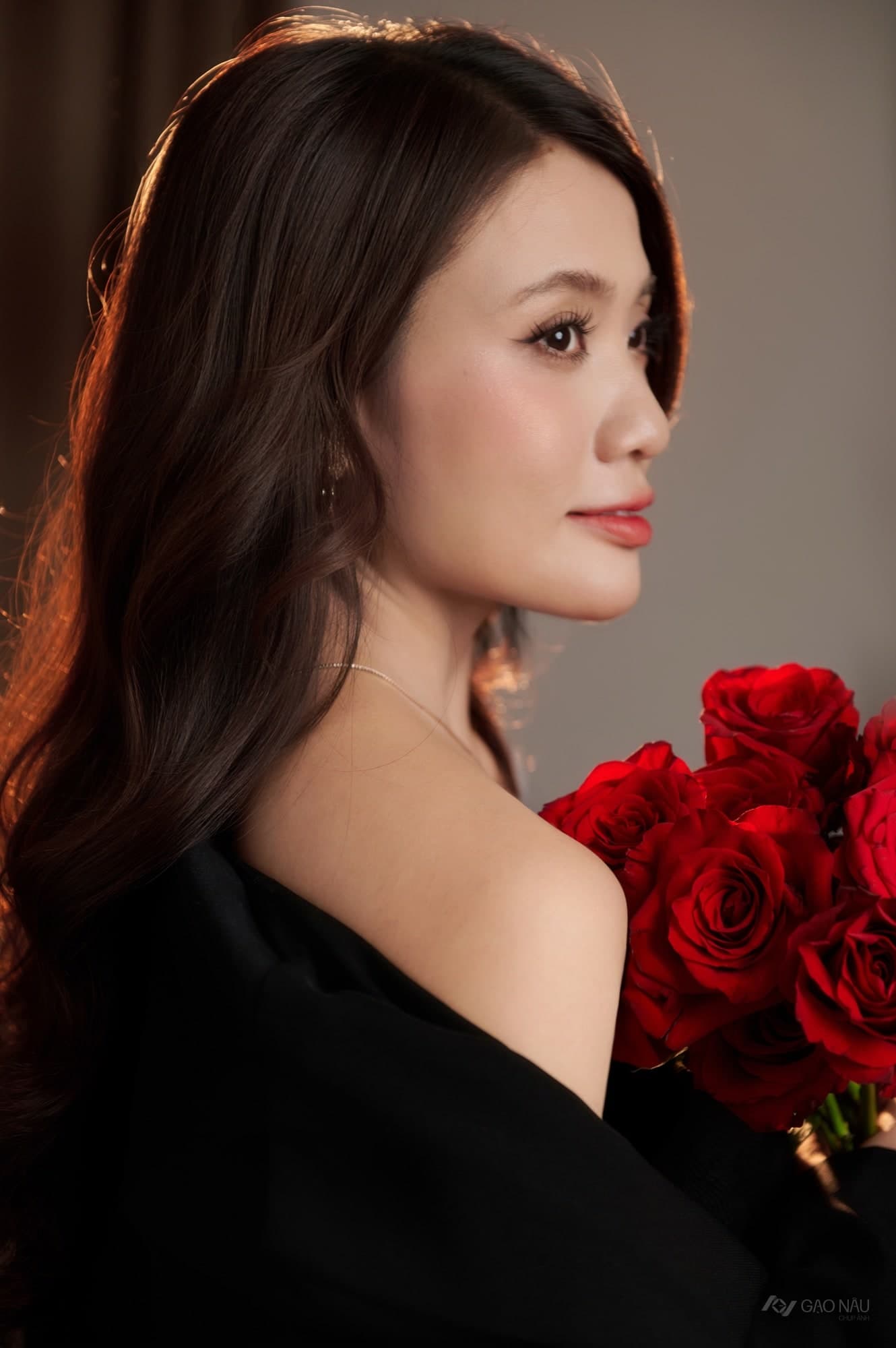 Cô ngồi nghiêng mặt về phía cửa sổ, hai tay ôm bó hoa hồng đỏ. Tóc búi gọn. Ánh sáng dịu, tông ấm, chất liệu phim điện ảnh.