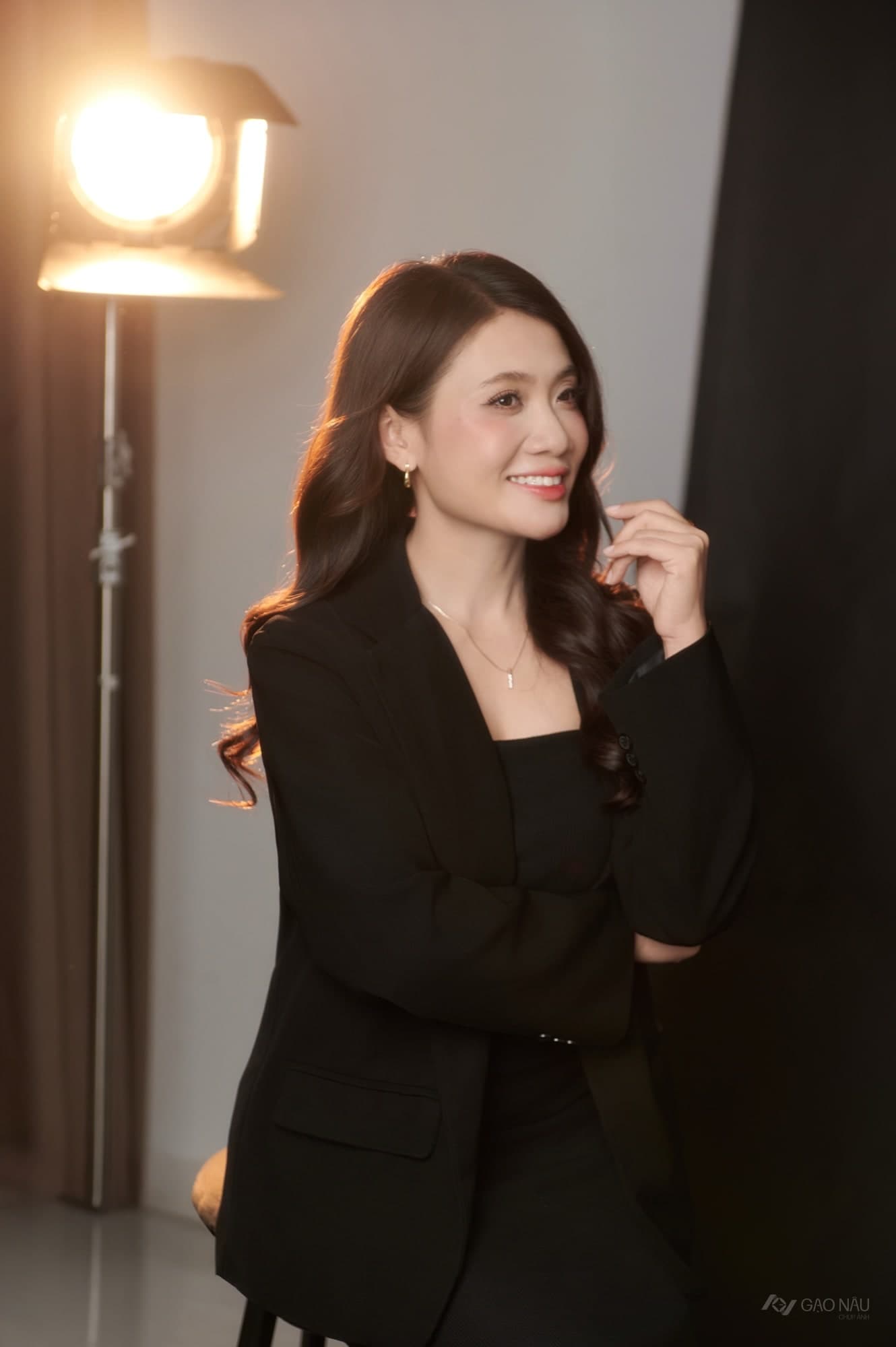 Người phụ nữ mặc suit đen, ngồi nghiêng, tay vuốt nhẹ mái tóc búi. Ánh đèn vàng ấm hắt từ bên phải. Ánh mắt nhìn xuống một điểm xa.