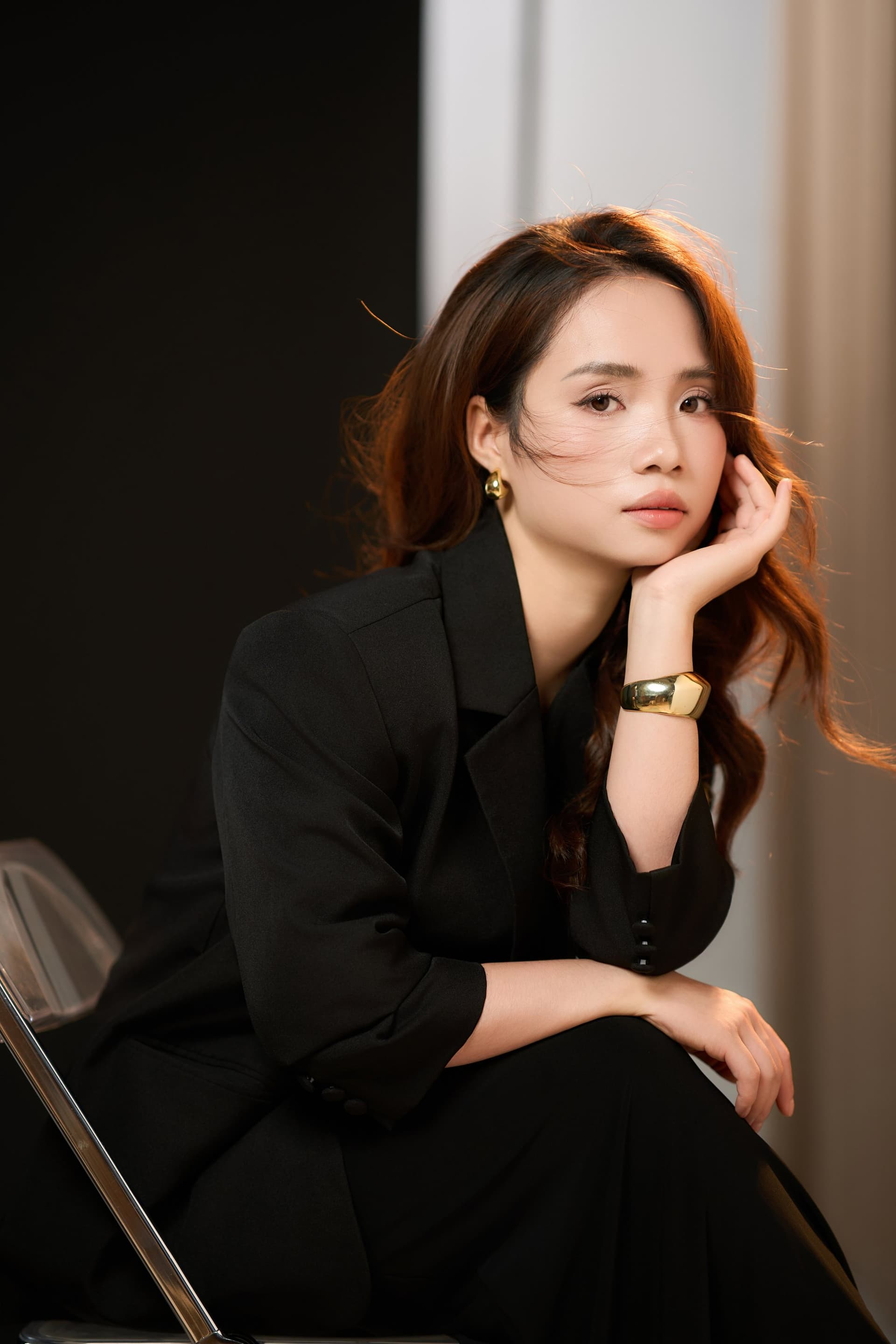 Cận người phụ nữ tay chống cằm, đeo vòng vàng, ánh nhìn trầm tư, blazer đen