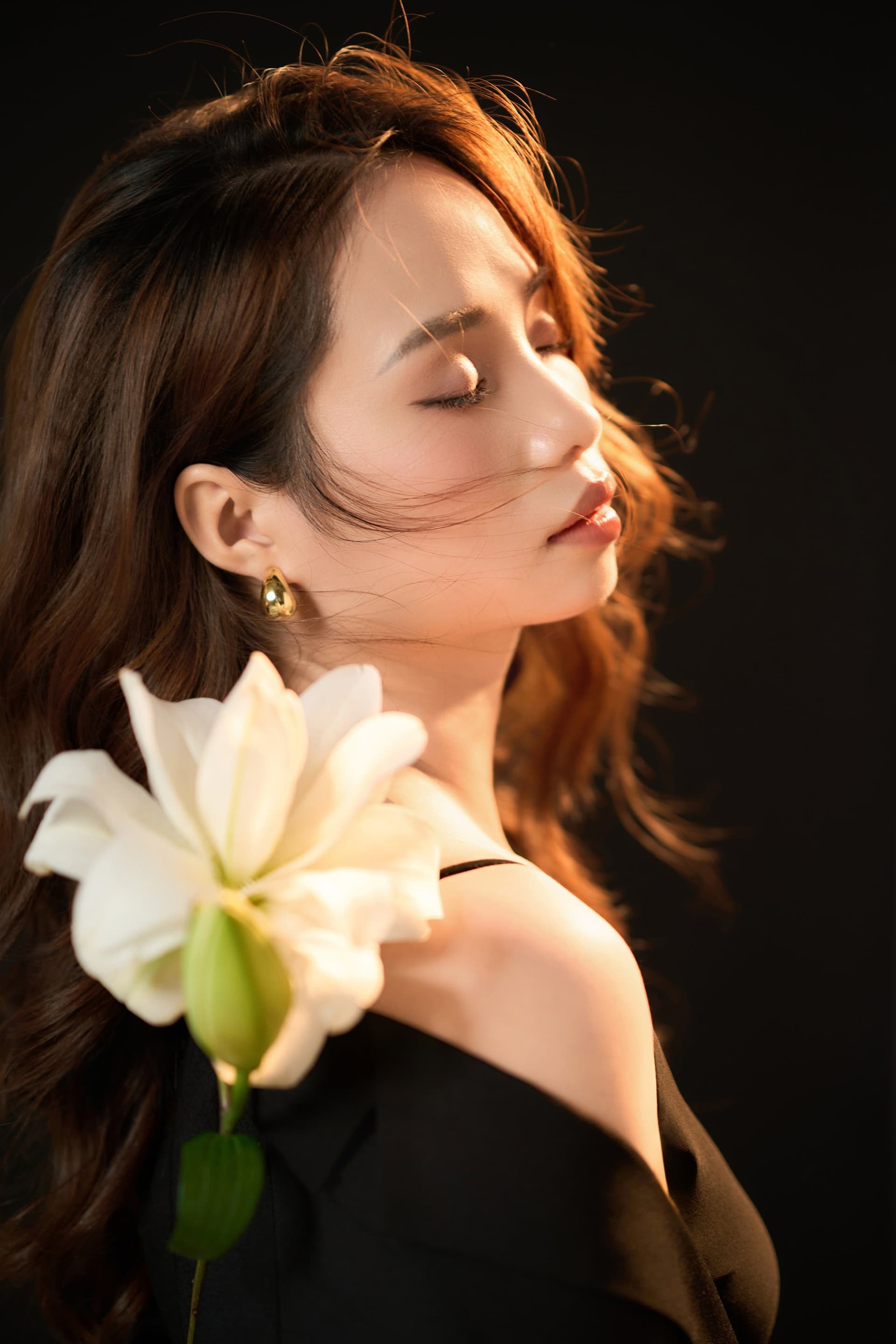 Cận mặt người phụ nữ mắt nhắm, một cánh loa kèn trắng đặt trên vai, biểu cảm lặng — đỉnh cảm xúc của bộ ảnh