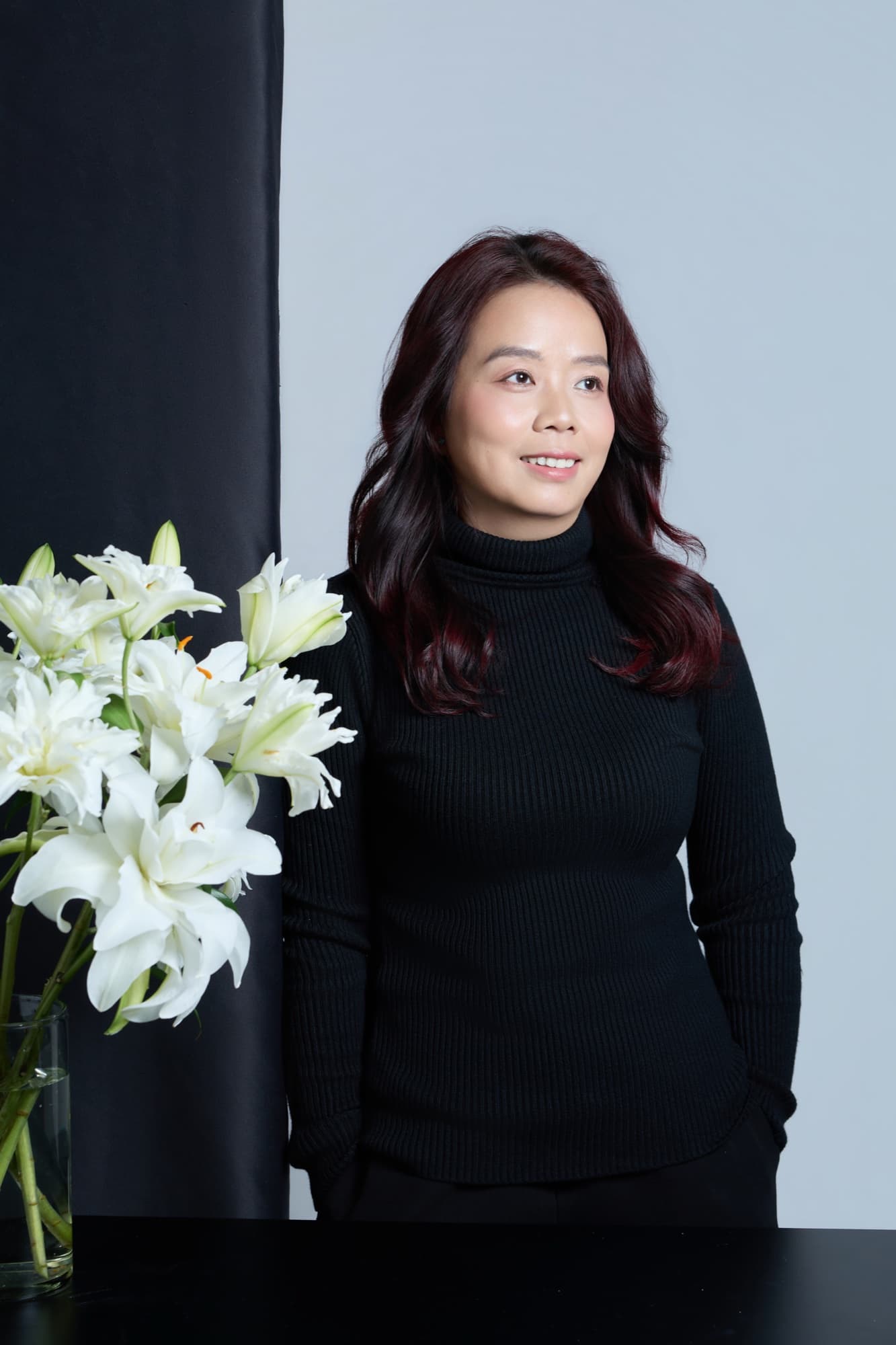 Người phụ nữ ngoài 40 mỉm cười nhẹ, đứng cạnh bình hoa loa kèn trắng, áo cao cổ đen, biểu cảm bình yên