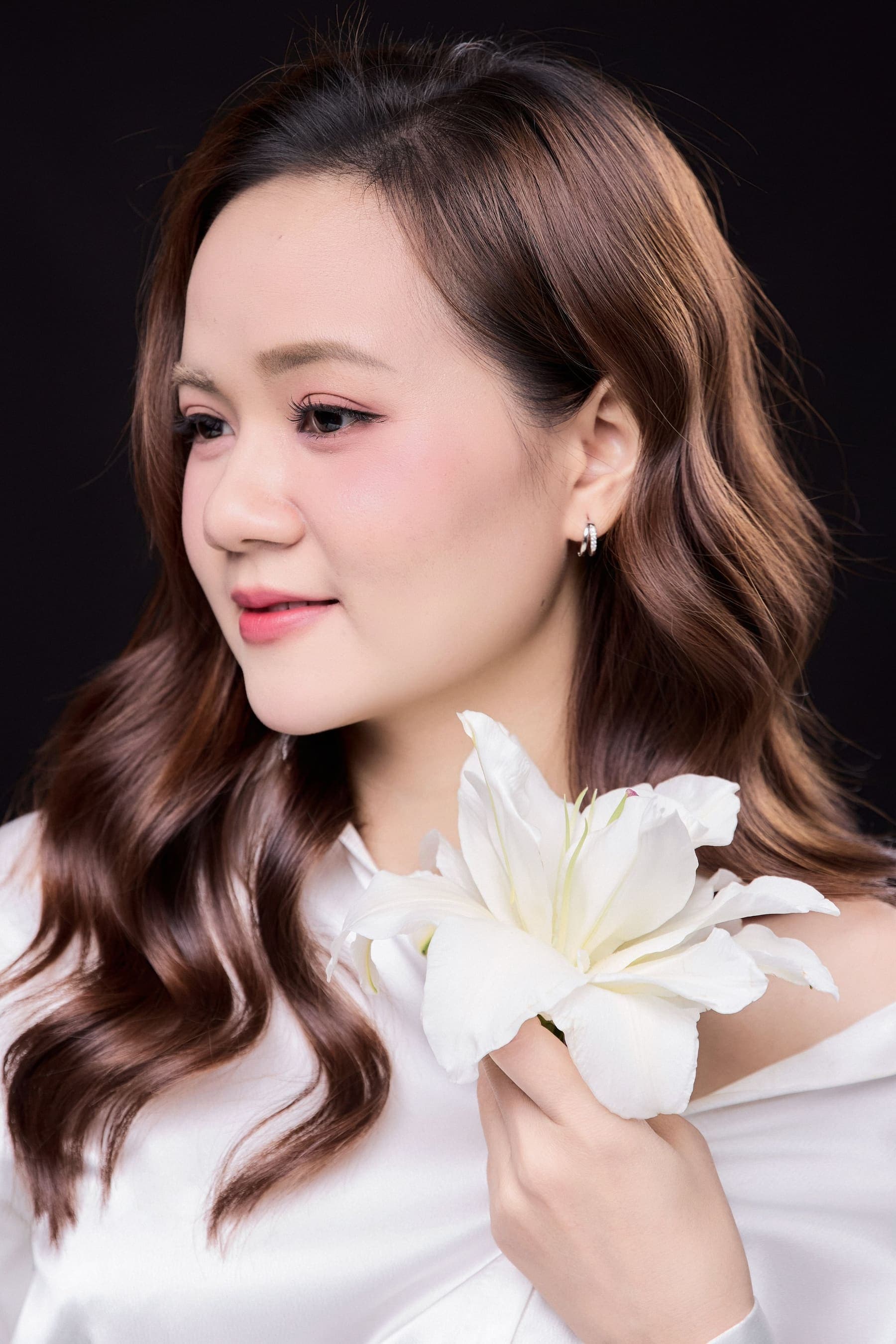 Thu Thảo ôm một bó hoa lily trắng, gương mặt sáng cười nhẹ — vẻ đẹp Bạch Ái: trắng, sạch, không cần nói nhiều