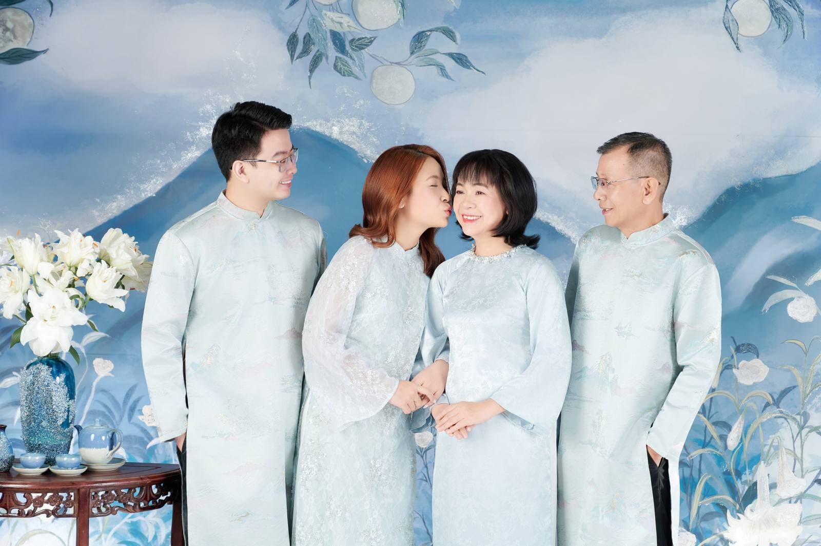 Gia đình bốn người mặc áo dài lụa xanh nhạt đứng cạnh nhau, background tranh núi mây, ánh sáng dịu