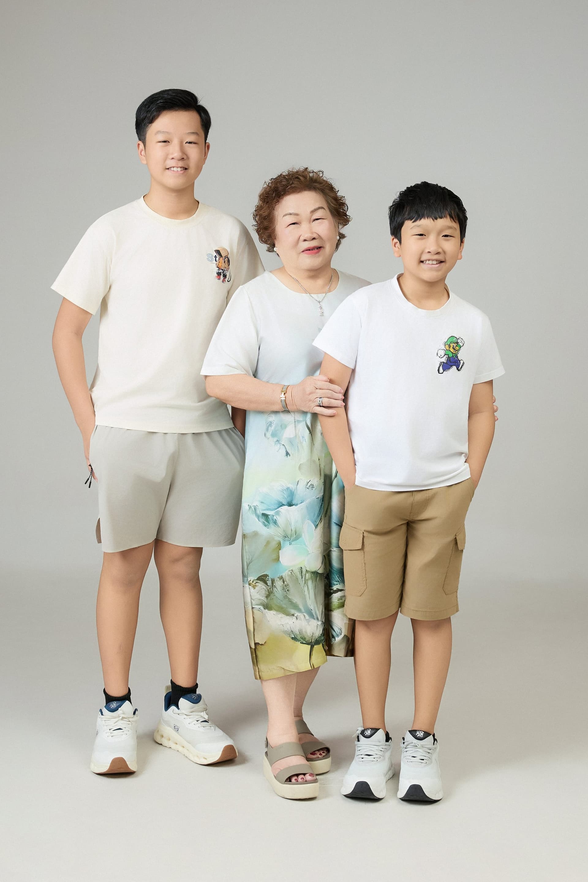 Năm người trong gia đình ba thế hệ — bà ngoại, bà nội, vợ chồng và con trai năm tuổi — đứng quây quần trong trang phục dịu, nền trắng