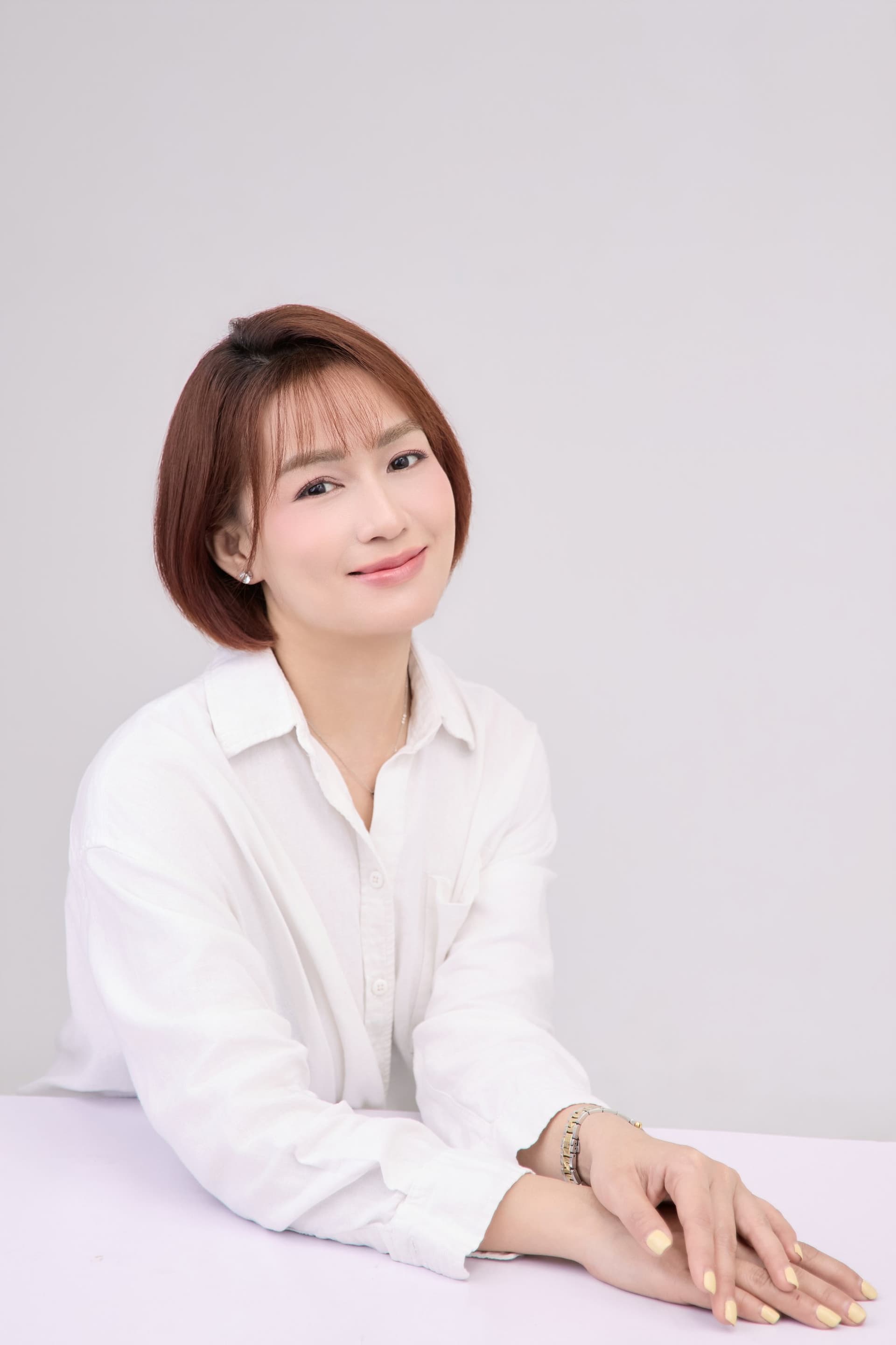 Chị tựa khuỷu tay lên bàn pink lavender, áo sơ mi trắng oversize, đồng hồ vàng-bạc trên cổ tay — lần đầu sau hai năm có thể ngồi yên
