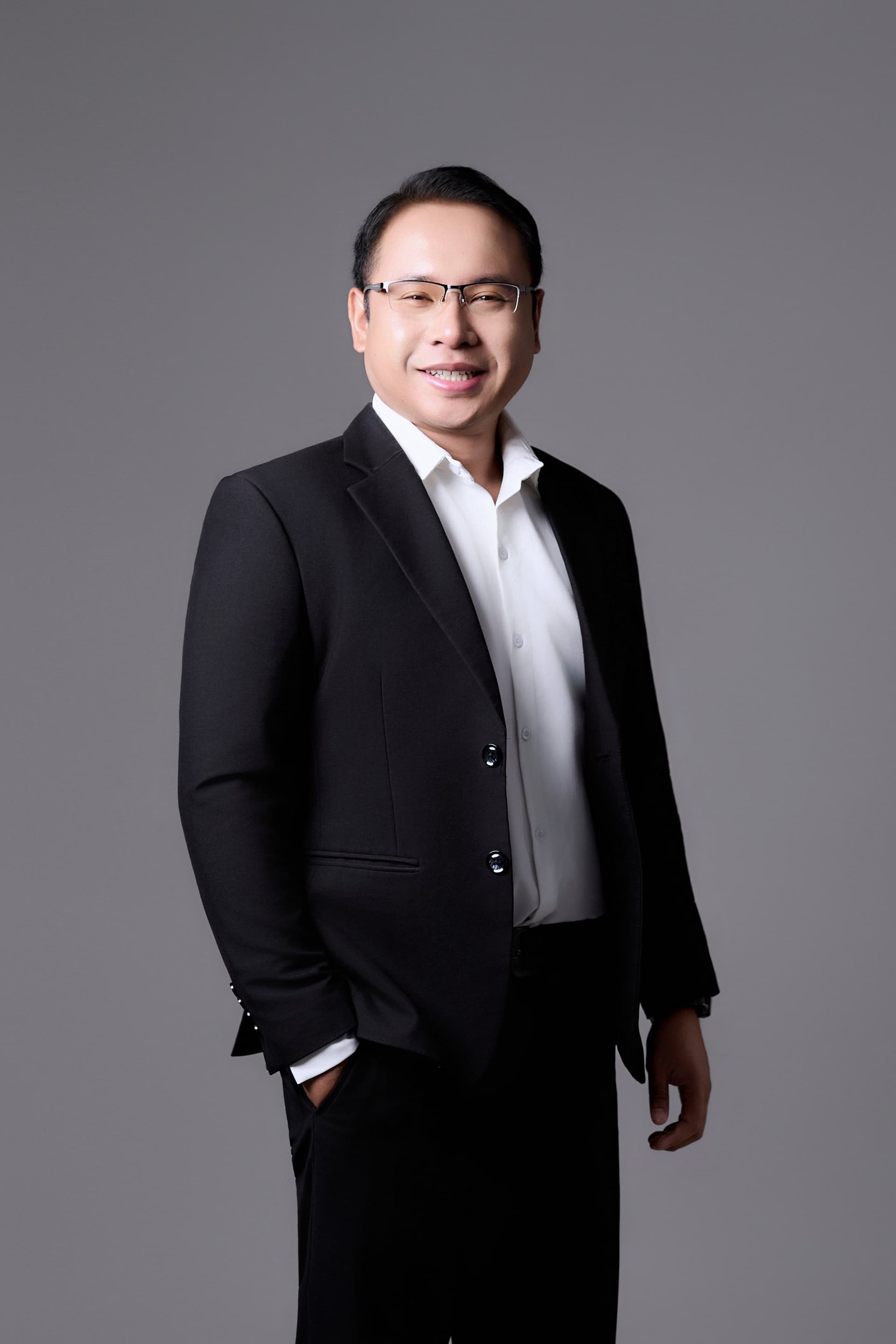 Người đàn ông vest đen, áo sơ mi trắng, mang kính, cười nhẹ — dáng đứng tay đút túi quần, ánh nhìn của một người làm học thuật lâu năm