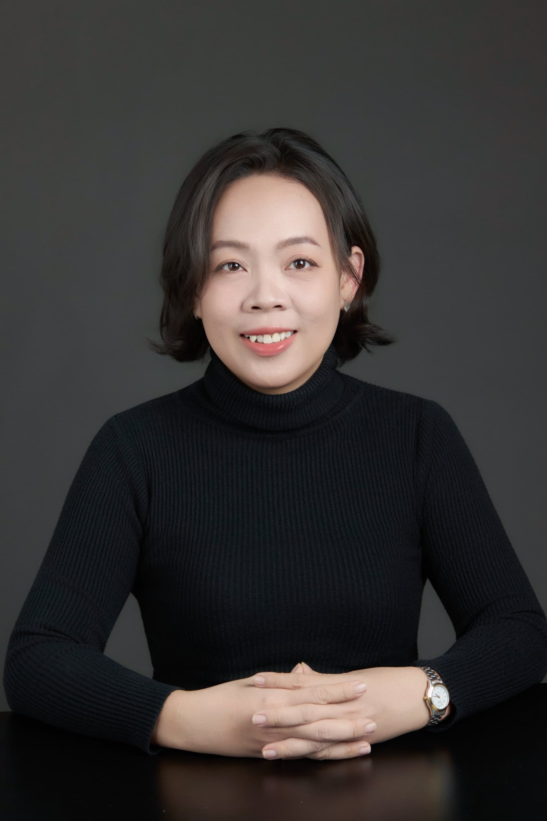 Bán thân — người phụ nữ tóc ngắn, áo cổ lọ đen, hai tay đan vào nhau đặt trên bàn, đeo đồng hồ kim loại trên cổ tay trái — tư thế của một người ngồi đối diện ai đó để lắng nghe