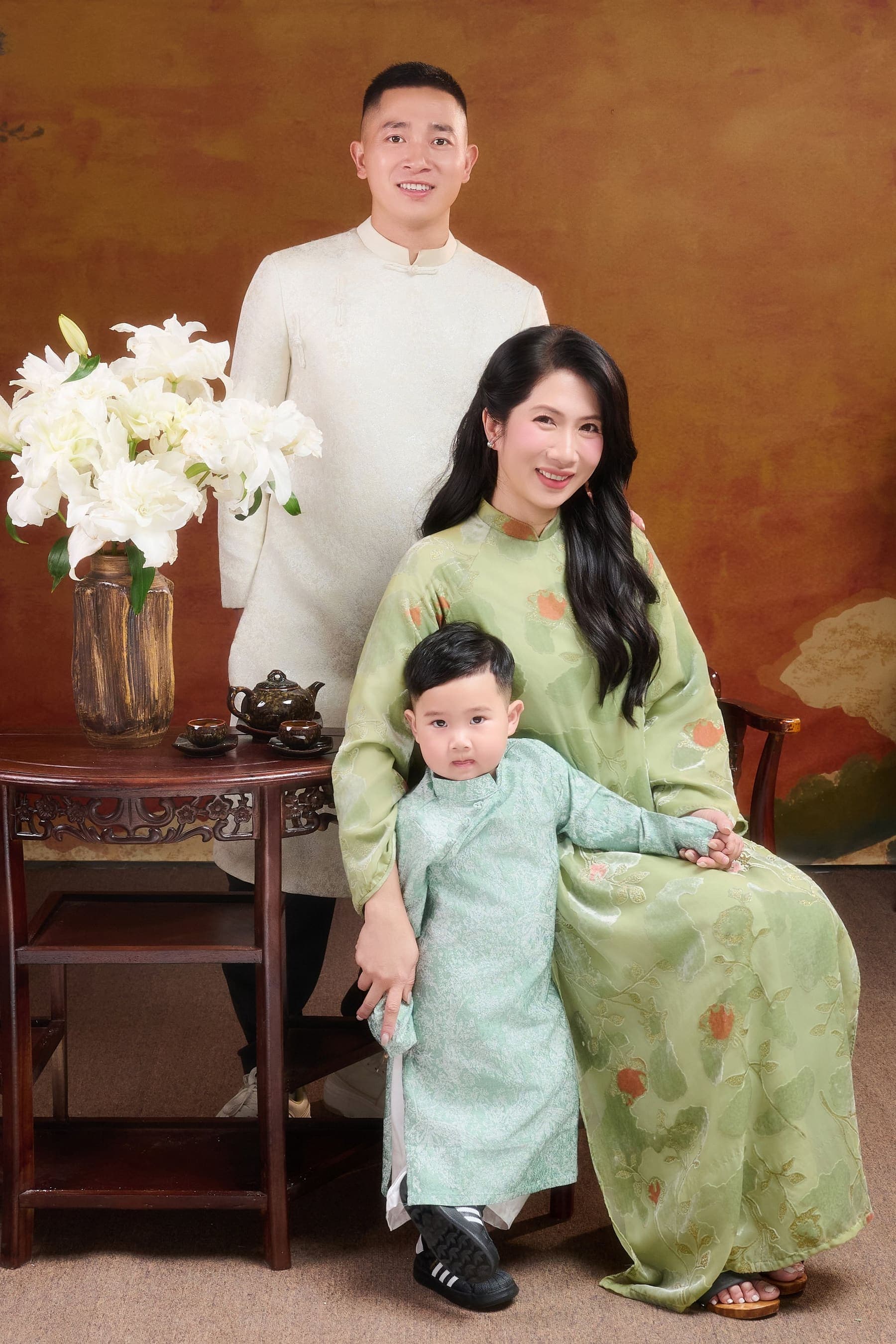 Cả nhà ngồi cùng nhau bên bình hoa loa kèn trắng — mẹ bầu, cha bế con nhỏ, một khung cảnh đời thường ấm áp