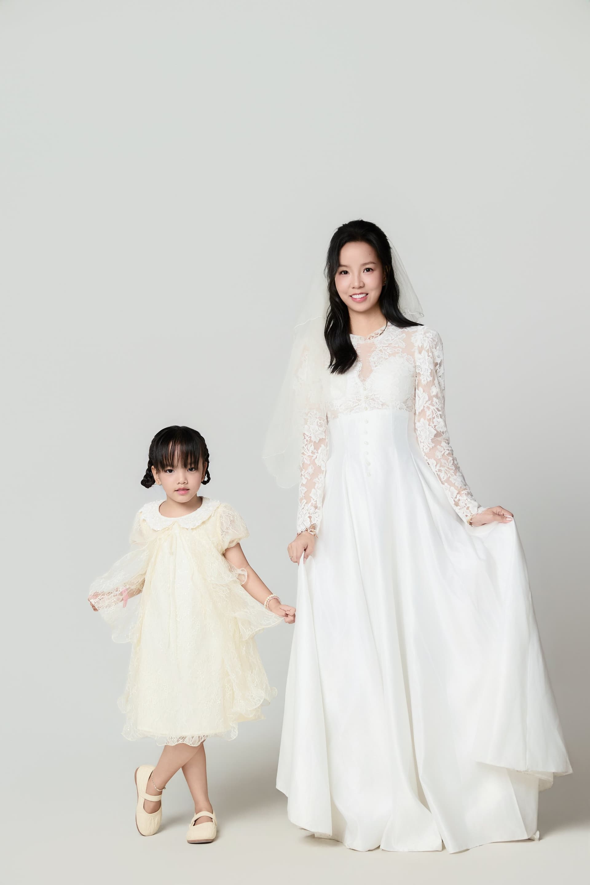 Người mẹ trong váy cô dâu trắng và con gái nhỏ váy trắng đứng đối diện nhau giữa nền dịu — hai vợ chồng cùng nhìn về một hướng, hướng đó là con