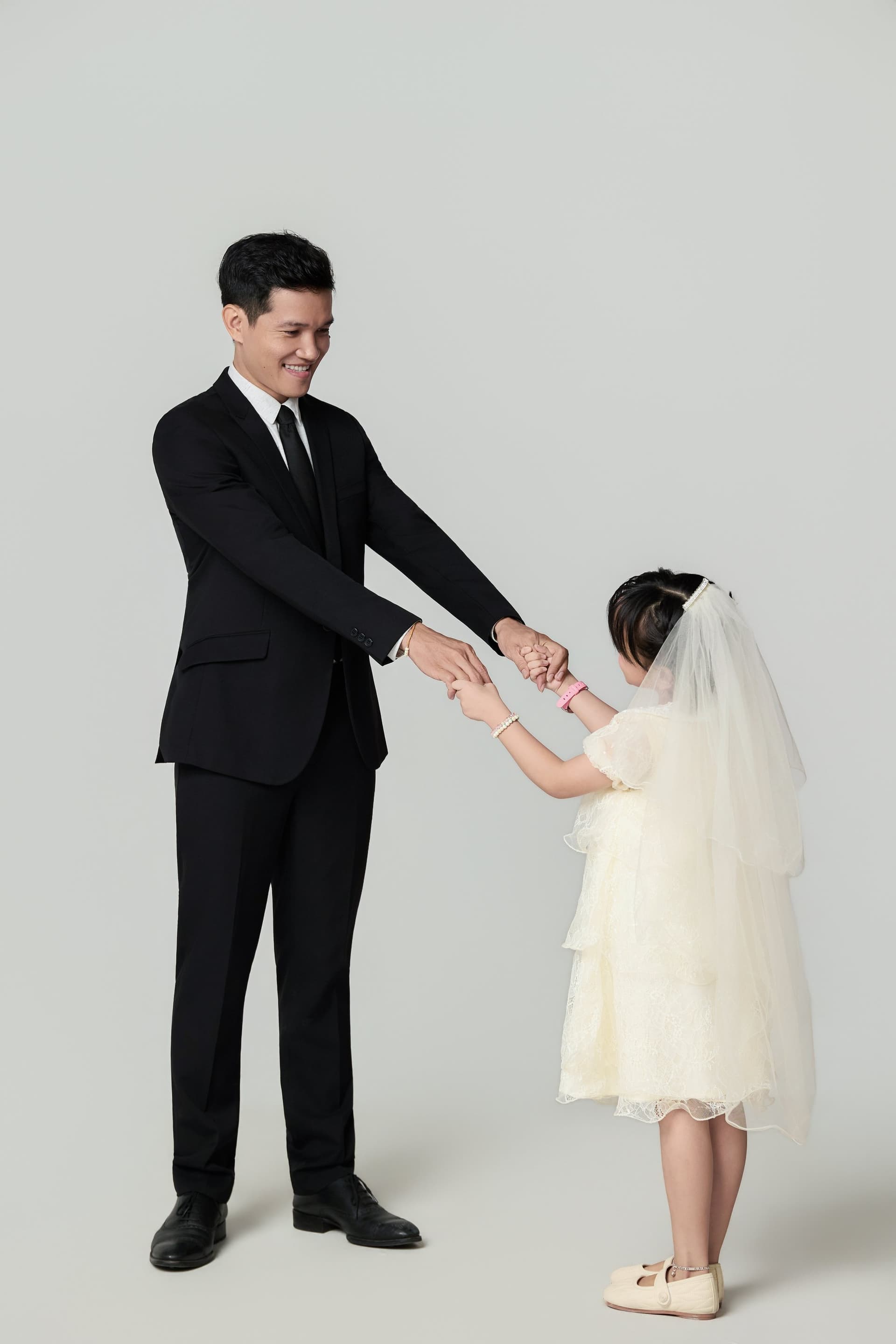 Người ba mặc vest đen cúi xuống nắm tay con gái nhỏ áo trắng — mỉm cười khi nhìn vợ và con