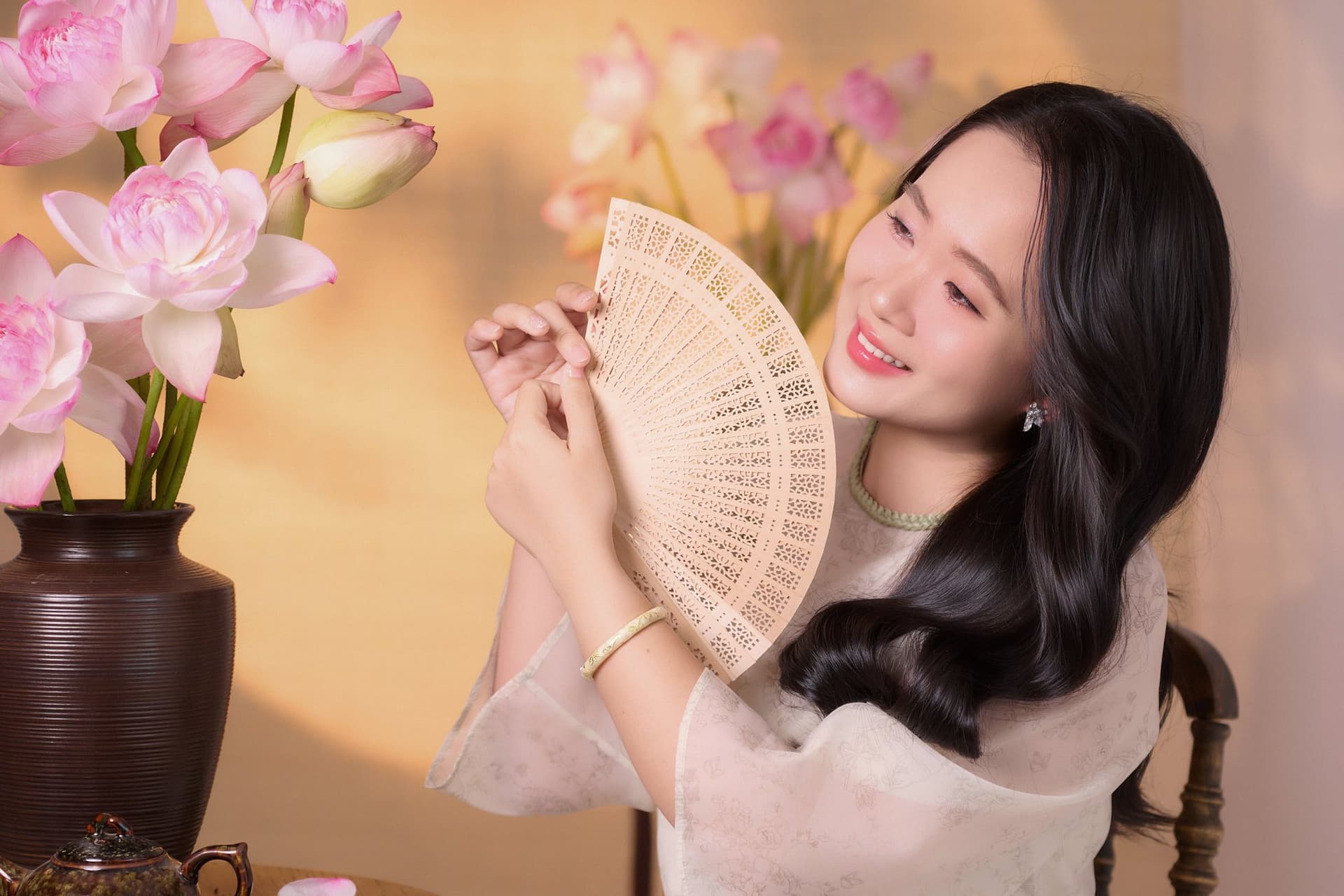 Người phụ nữ áo trắng cười dịu, tay cầm quạt giấy, nền tone vàng be ấm