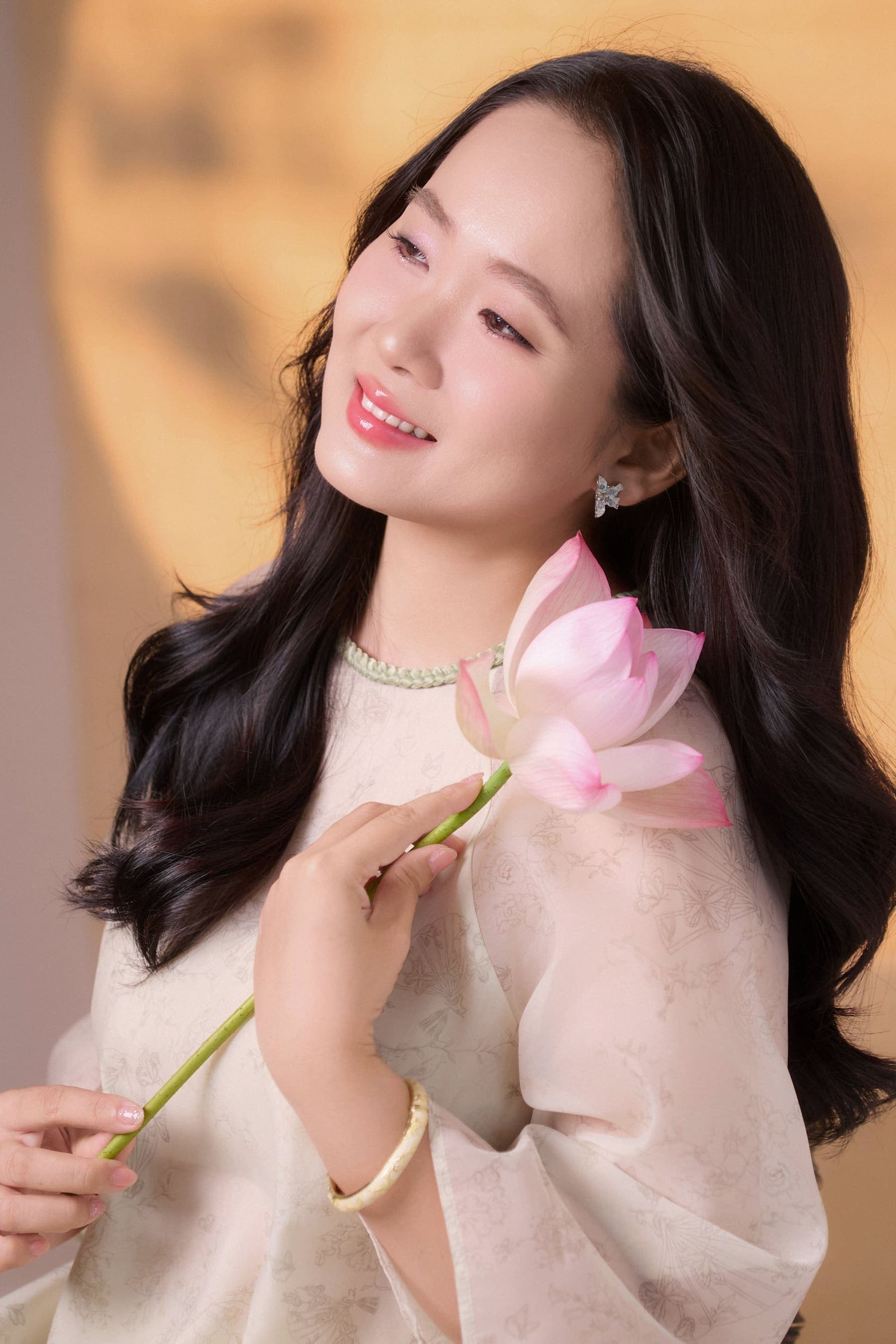 Người phụ nữ ngồi cười dịu, tay đỡ bông sen, nền hoa sen tone ấm