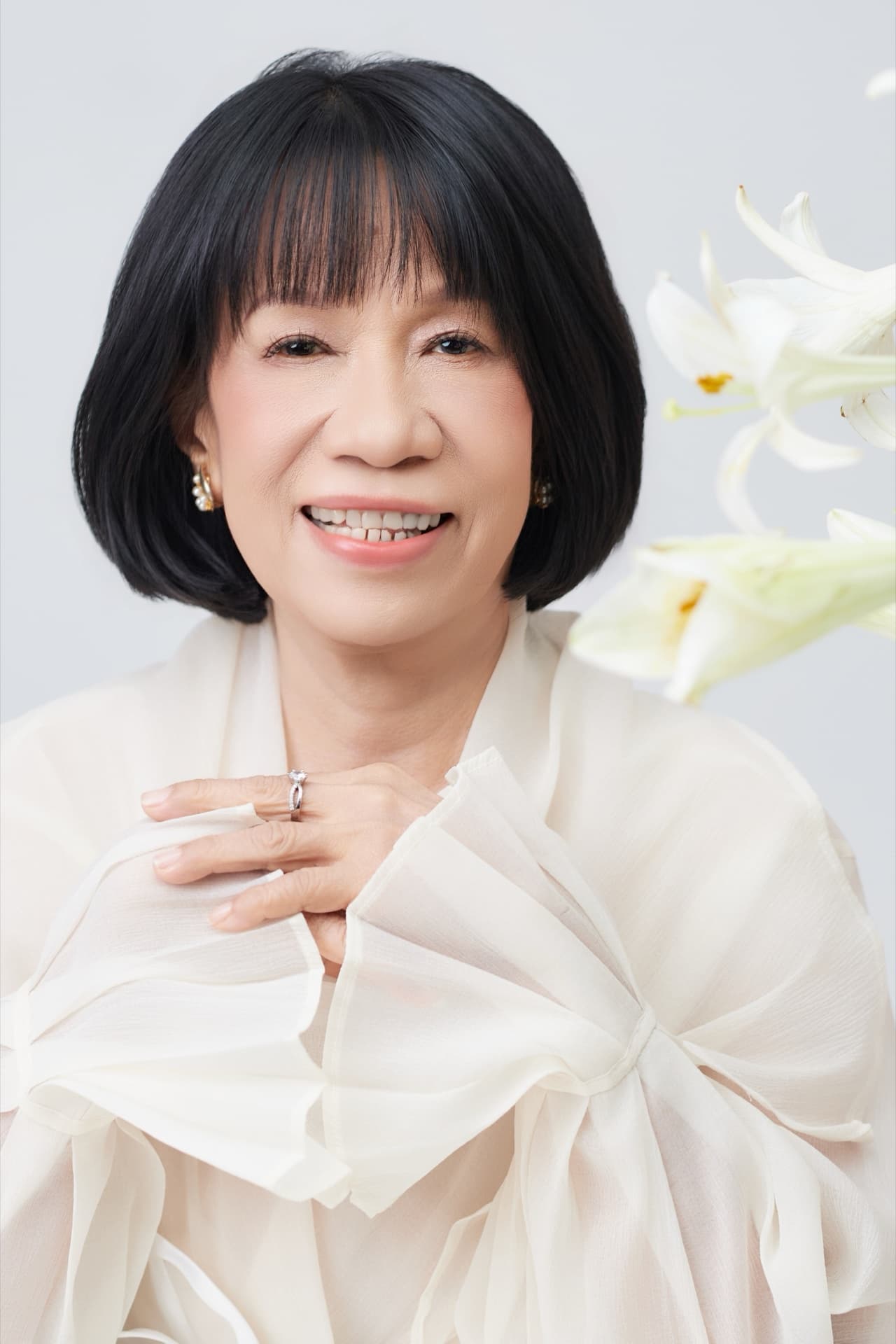 Cô Nguyệt Mai 66 tuổi, áo trắng, tay ôm bó loa kèn trắng, ánh sáng tự nhiên dịu, khung mặt hiền hậu