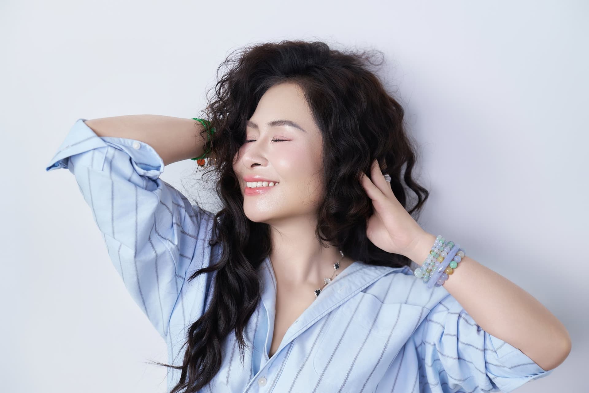 Người phụ nữ tóc xoăn dài, cười nhẹ, tay sau gáy — dáng của người đang biết lại chính mình