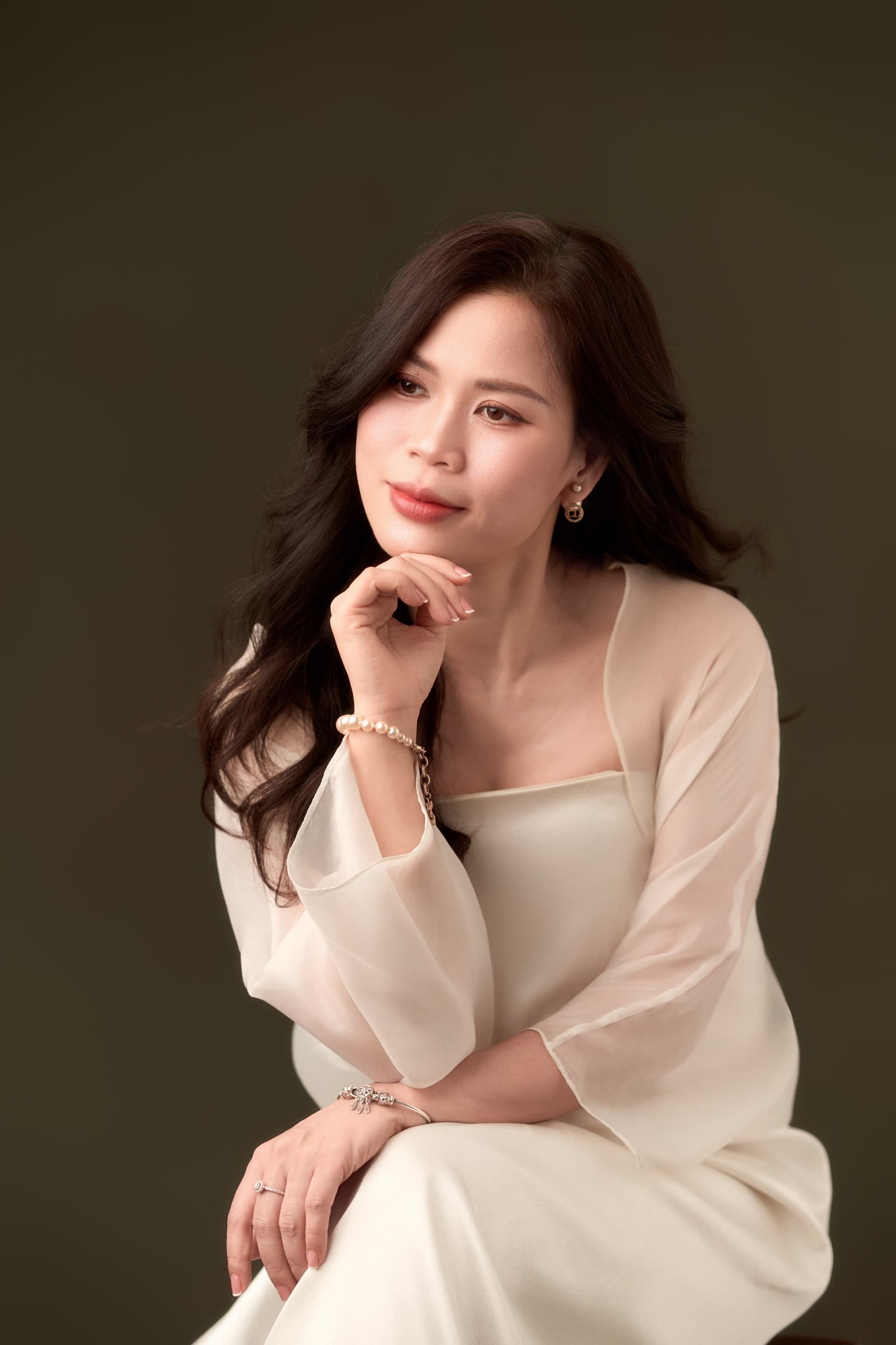 Người phụ nữ áo trắng tóc đen buông dài, bàn tay khẽ chạm cằm, ánh mắt nhìn xa — khoảnh khắc lặng giữa đời sống bận của một dáng người vốn năng động