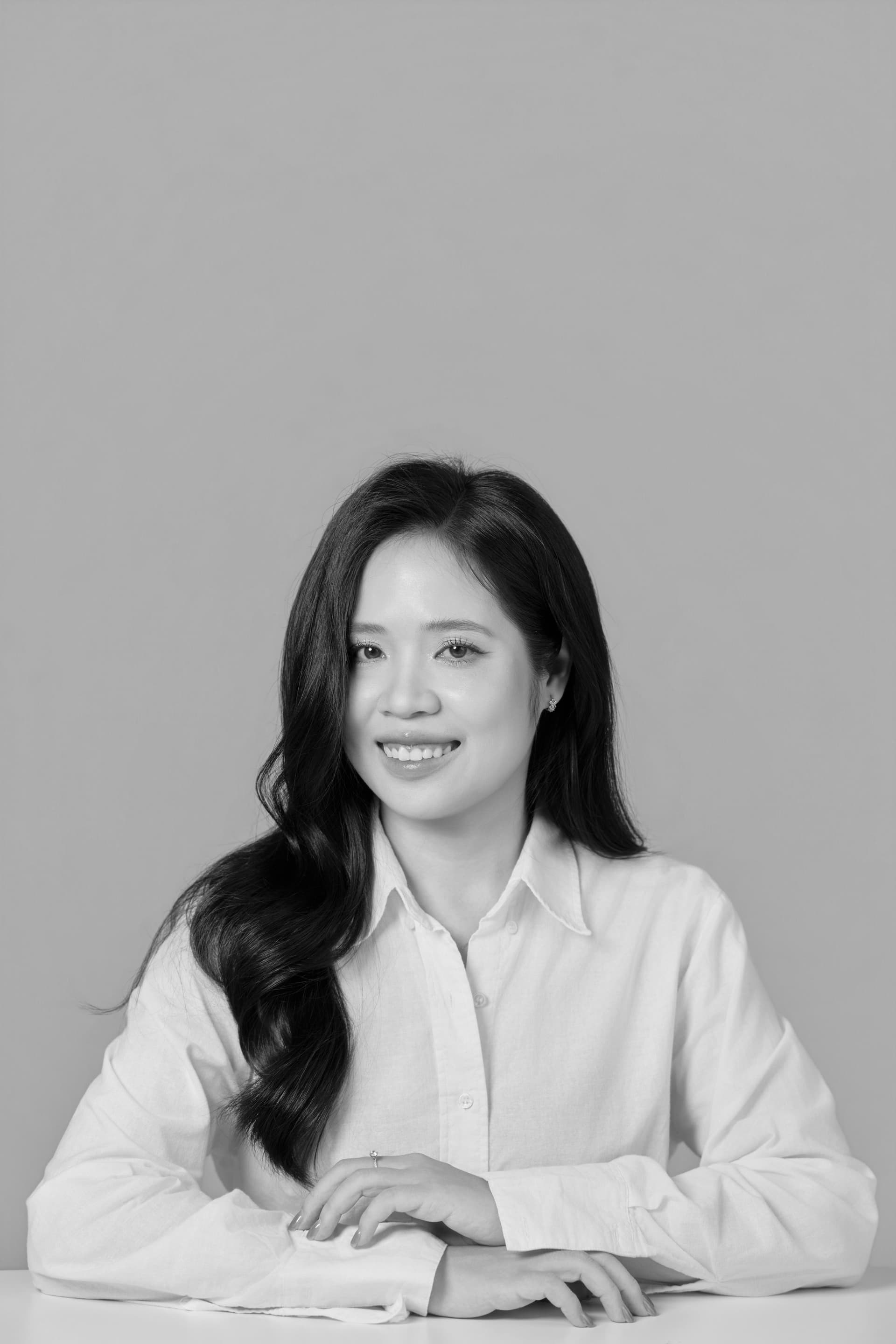 Người phụ nữ áo sơ mi trắng ngồi bên bàn, hai tay đan lại, đôi mắt nhìn xa xăm — khung đen trắng trầm tư, gợi những năm tháng tự nghiêm khắc với chính mình.