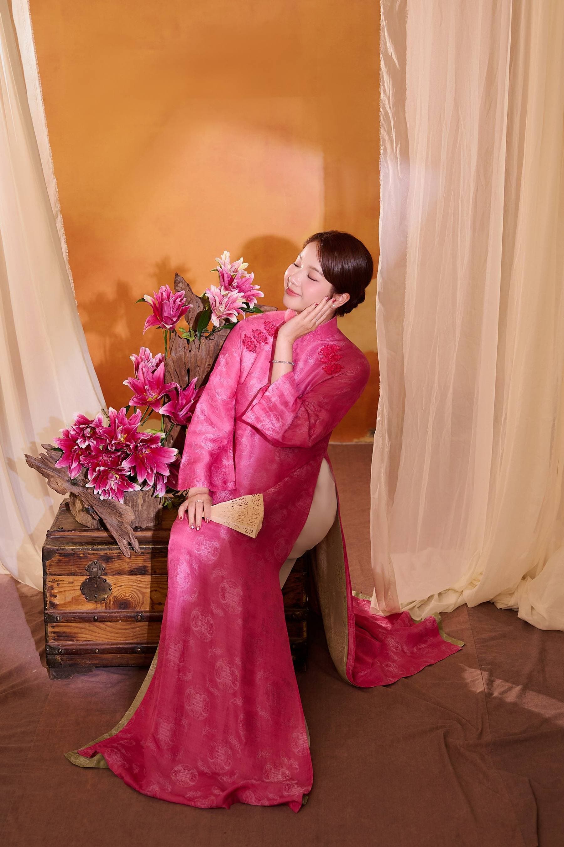 Chị Thanh đứng giữa nền hoa hồng cam, áo dài hồng — toàn cảnh trong khoảnh khắc soi gương