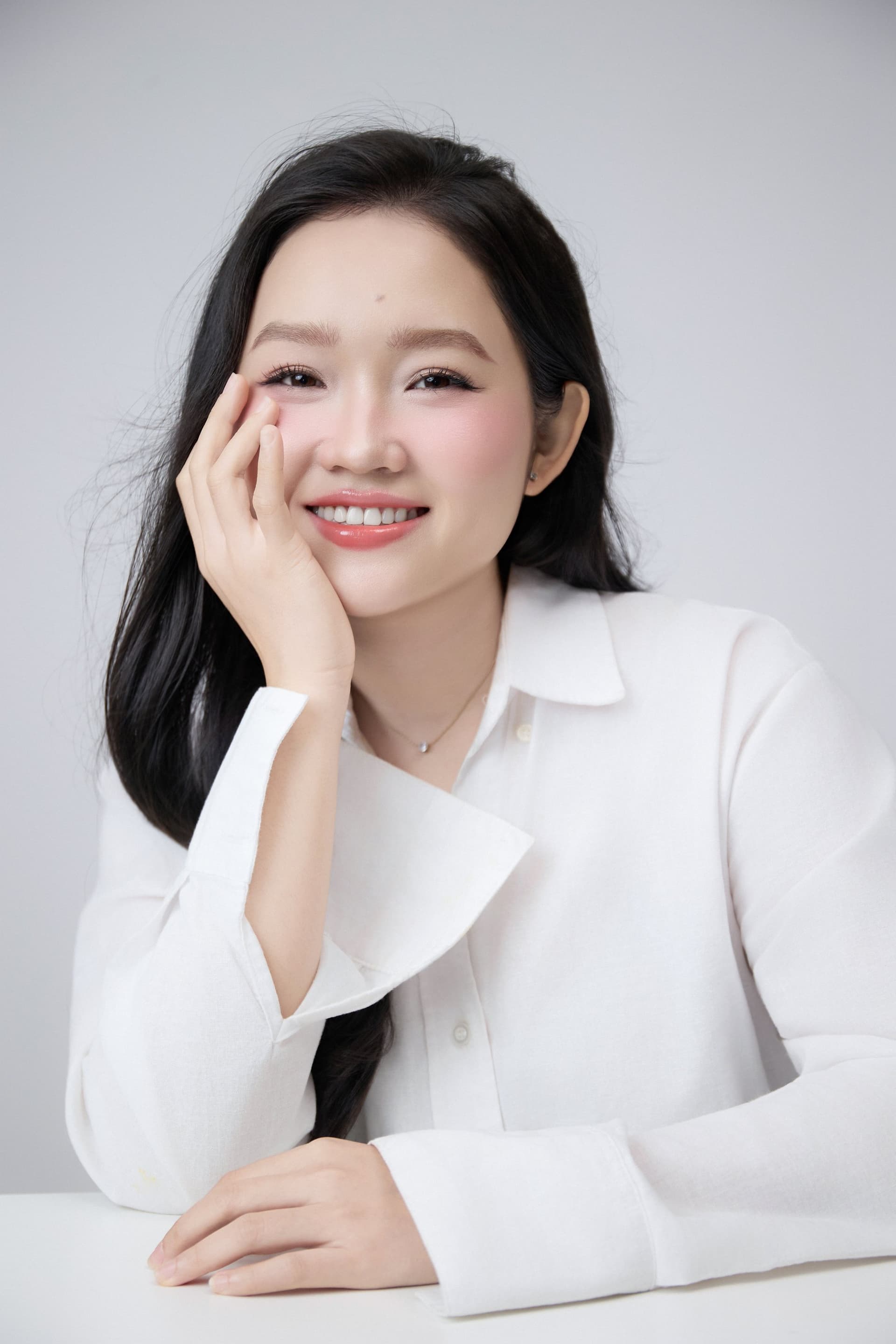 Cô gái áo trắng cười nhẹ, tay đỡ má, ánh mắt sáng, gương mặt còn nét trẻ trung của tuổi hai mươi tám