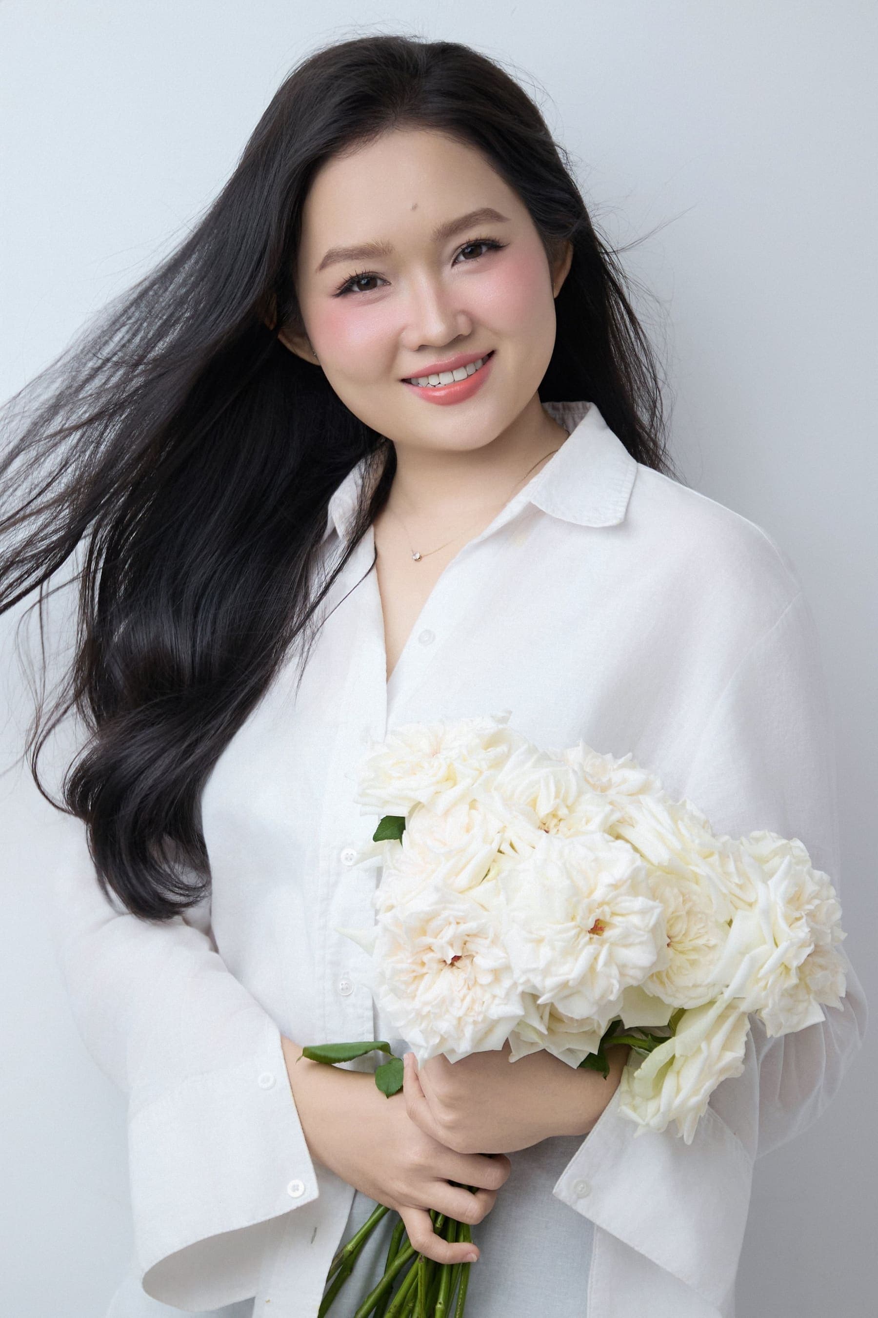 Phương Thảo ôm bó hoa hồng trắng cuống dài, mặc áo sơ mi trắng, cười nhẹ — một khoảnh khắc giữ lại tuổi hai mươi tám