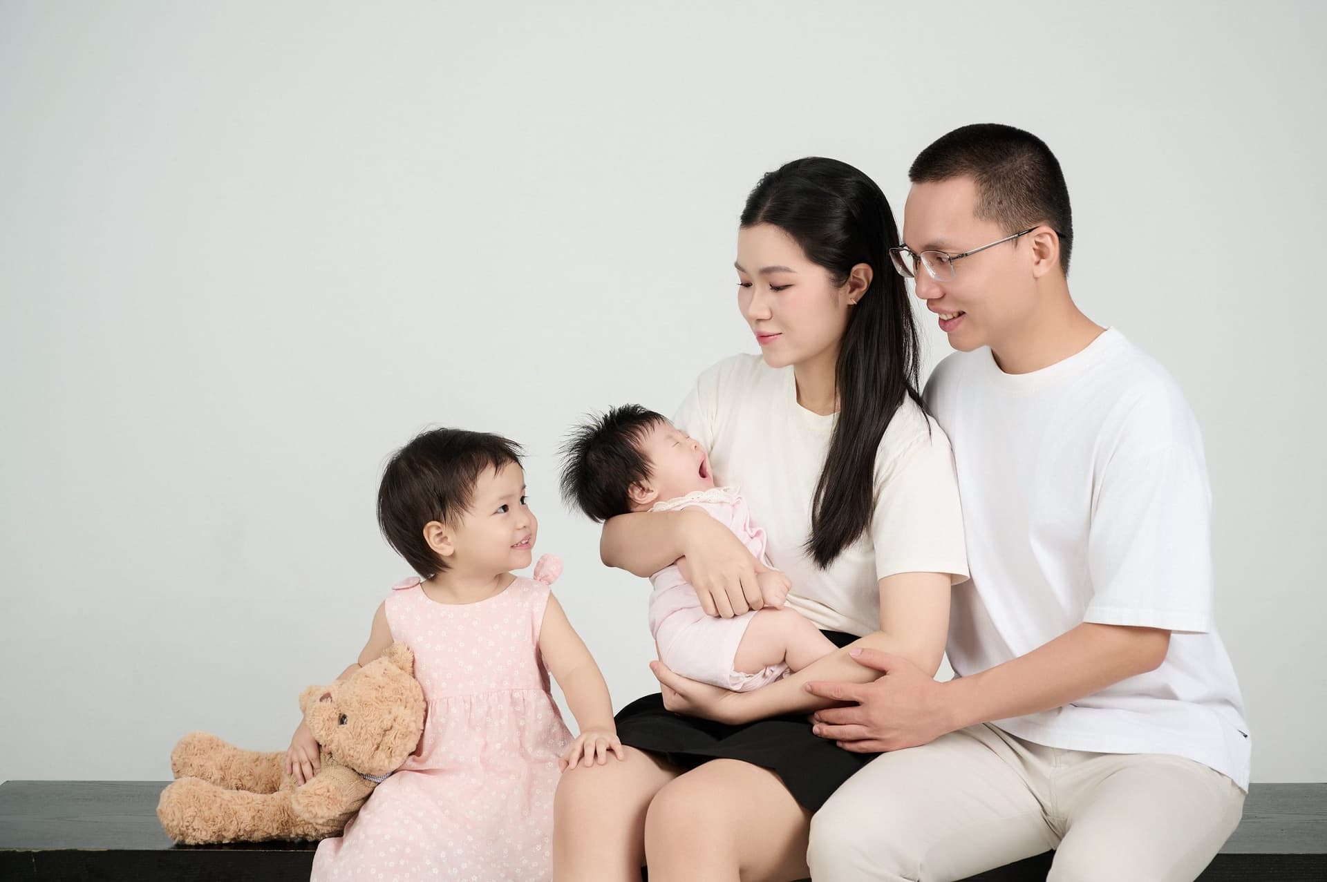 Ba mẹ cùng hai con ngồi quây quần trên sàn, ba ôm bé lớn cười rạng rỡ bên cạnh mẹ bế em nhỏ