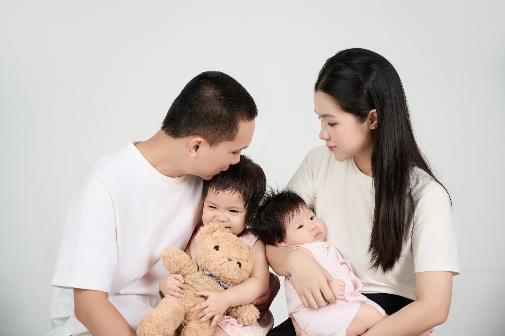Cả nhà cùng cúi đầu cười hạnh phúc, ngắm em bé sơ sinh nằm ngoan bên cạnh chú gấu bông nhỏ trên thảm