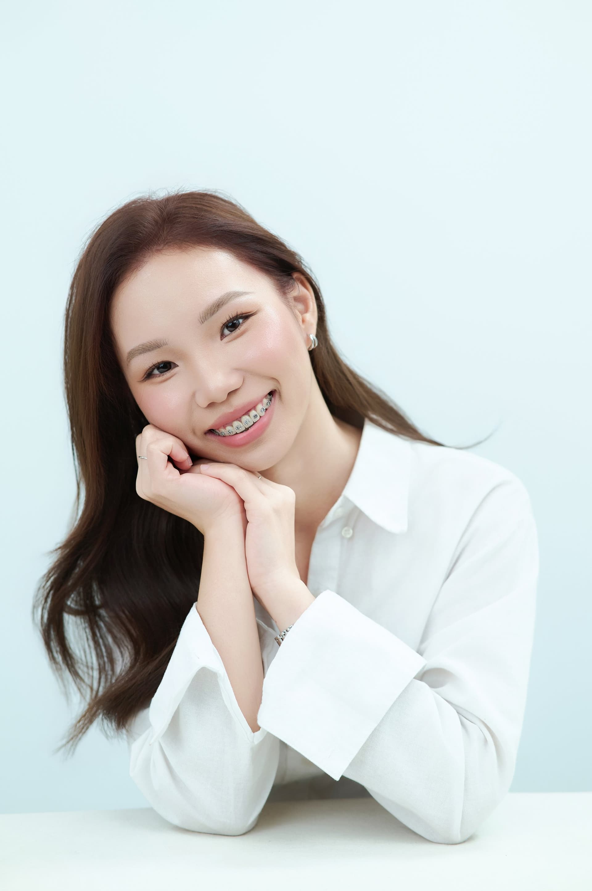 Người phụ nữ hai tay đỡ cằm, cười dịu, áo sơ mi trắng cổ bẻ — chân dung bán thân
