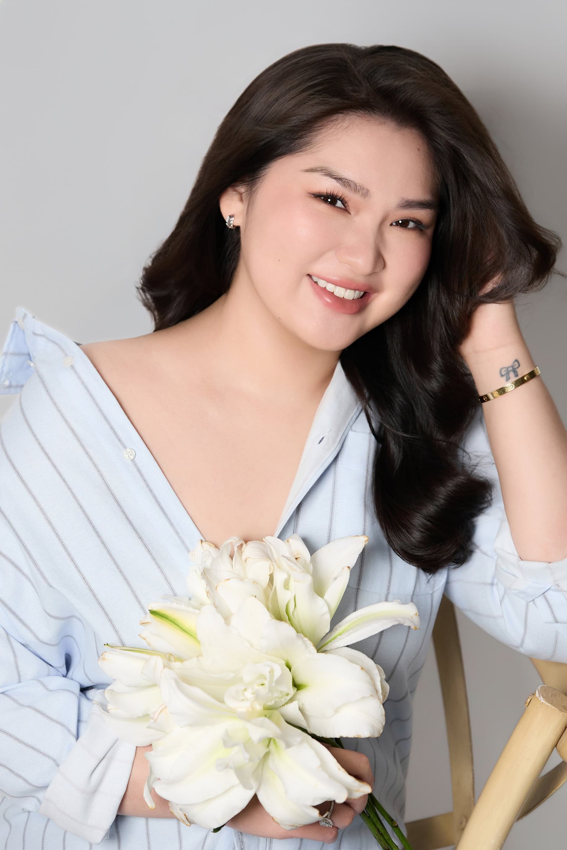 Người phụ nữ đứng mỉm cười dịu dàng với bàn tay khẽ chạm cằm trước nền trắng