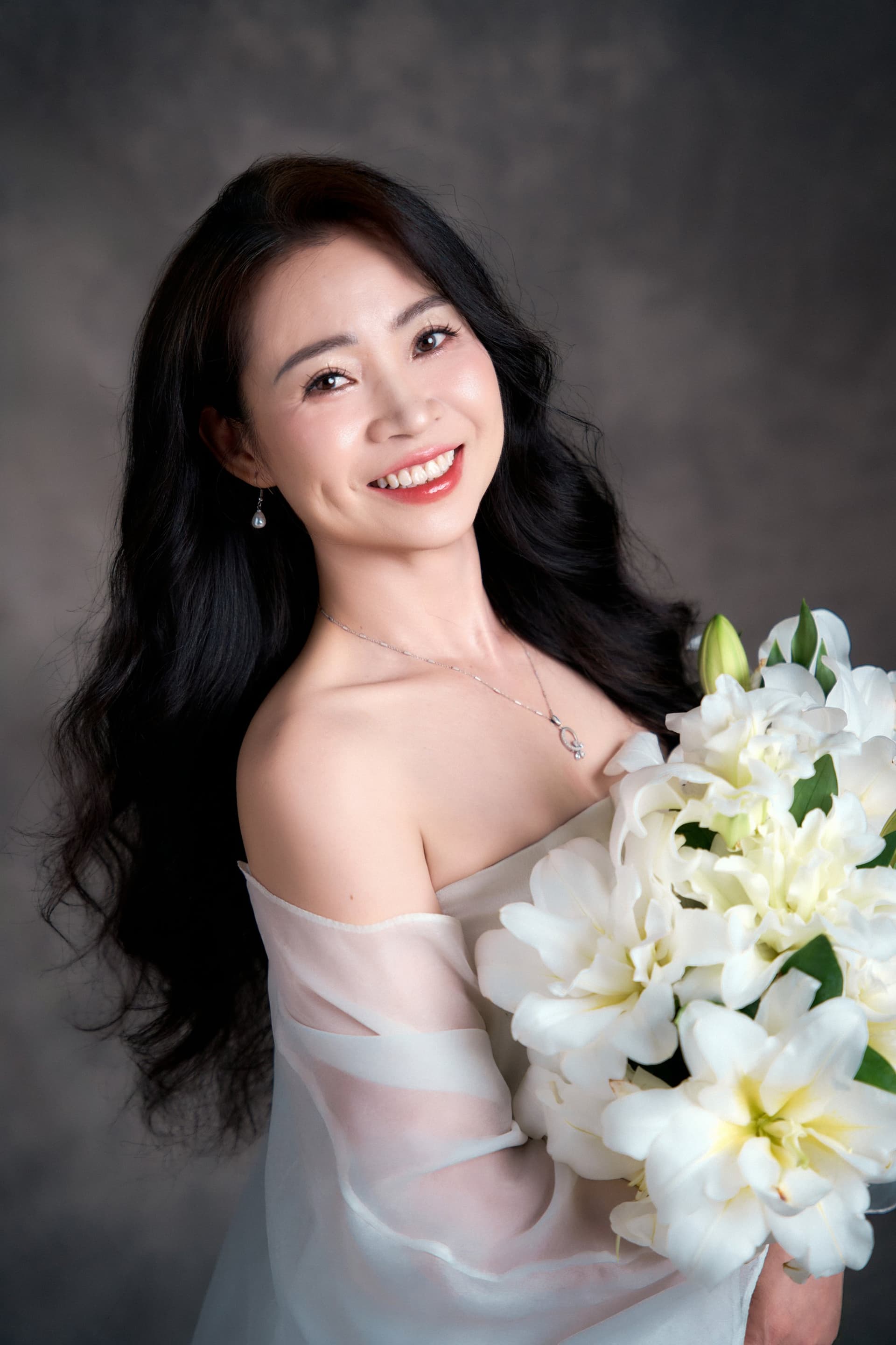 Cô Mận với góc nhìn nghiêng ôm bó hoa ly trắng nở rộ, khẽ cúi đầu mỉm cười nhẹ nhàng đầy bình yên