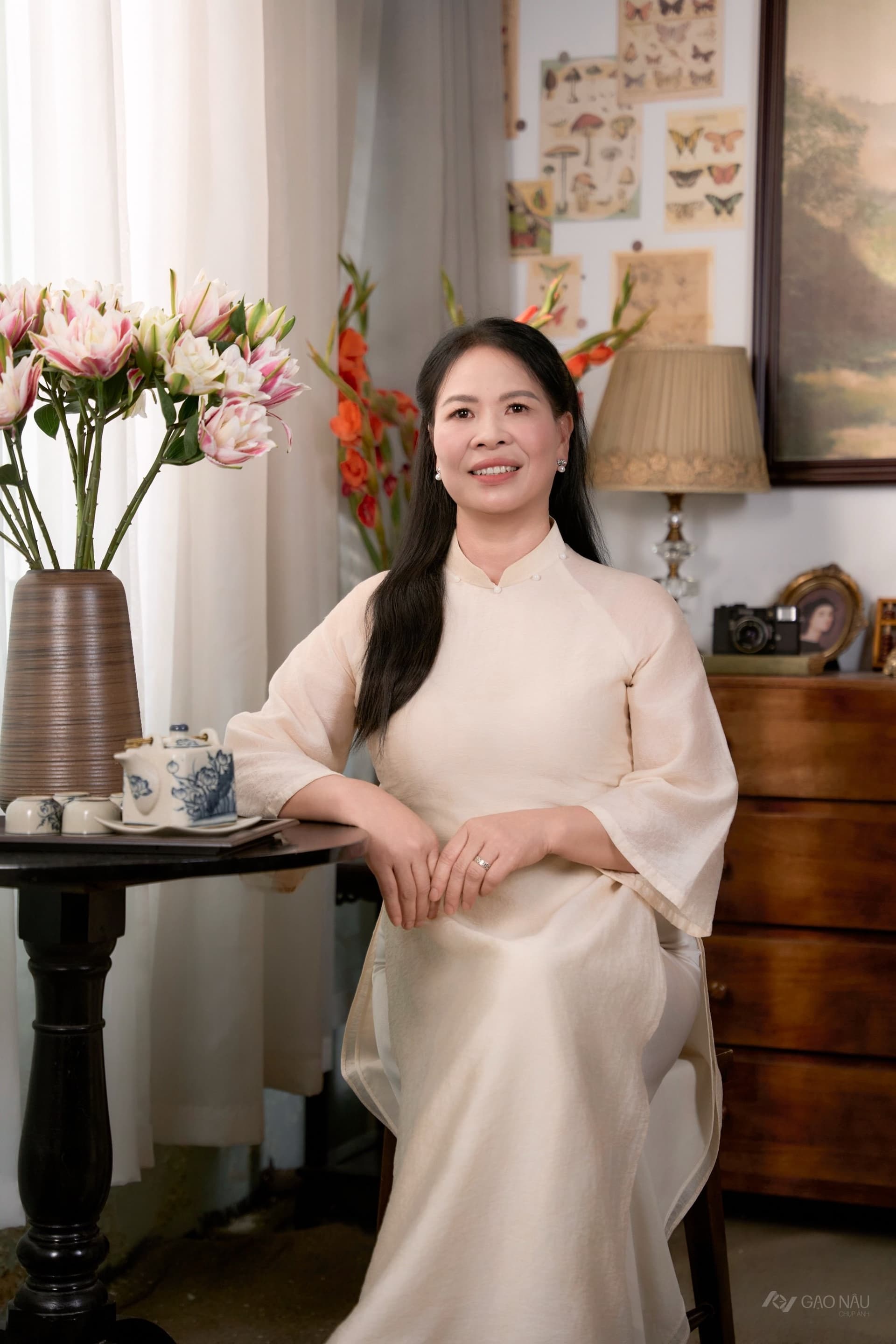 Người phụ nữ trung niên mặc áo dài lụa màu kem, ngồi yên bên bàn nhỏ có bình hoa loa kèn trắng, ánh sáng tự nhiên chiếu nhẹ