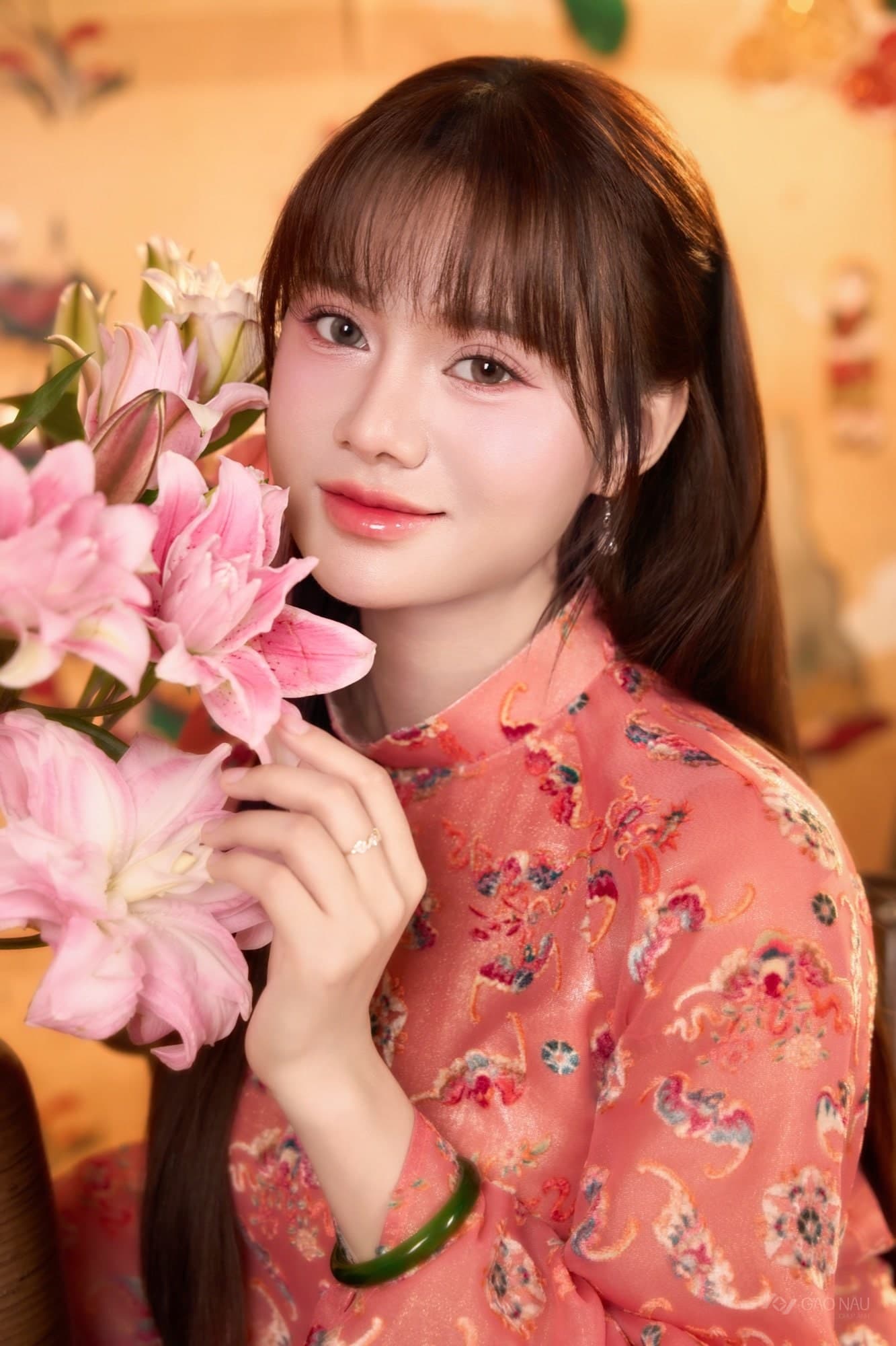 Tiểu Ngáo mặc áo dài đỏ hoa văn Tết, tay cầm bó hoa lily hồng, cười dịu