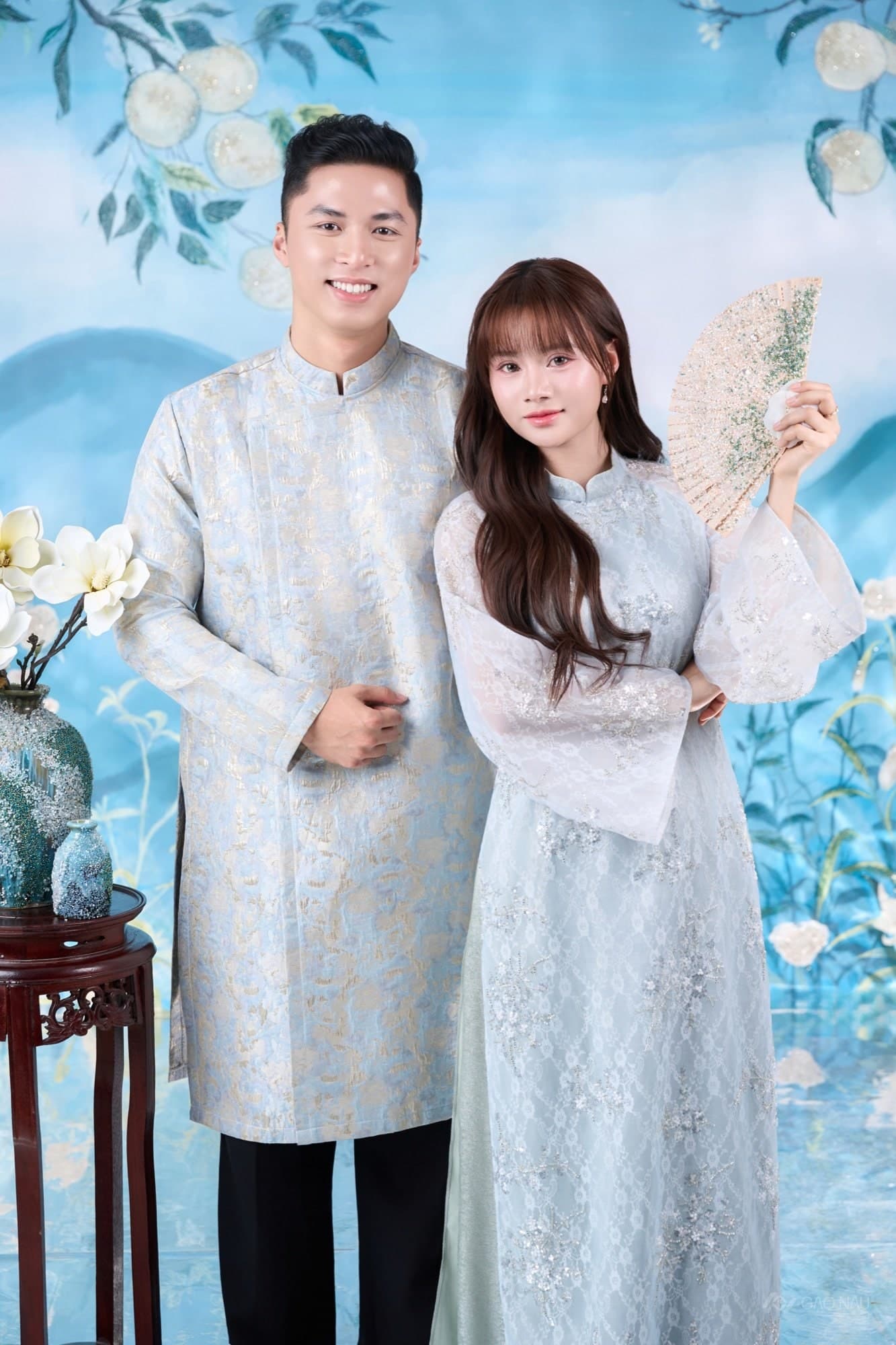 Tiểu Ngáo và Hoàng Hôn mặc áo dài xanh ngọc, cô cầm quạt giấy, phông hoa sen nền xanh pastel