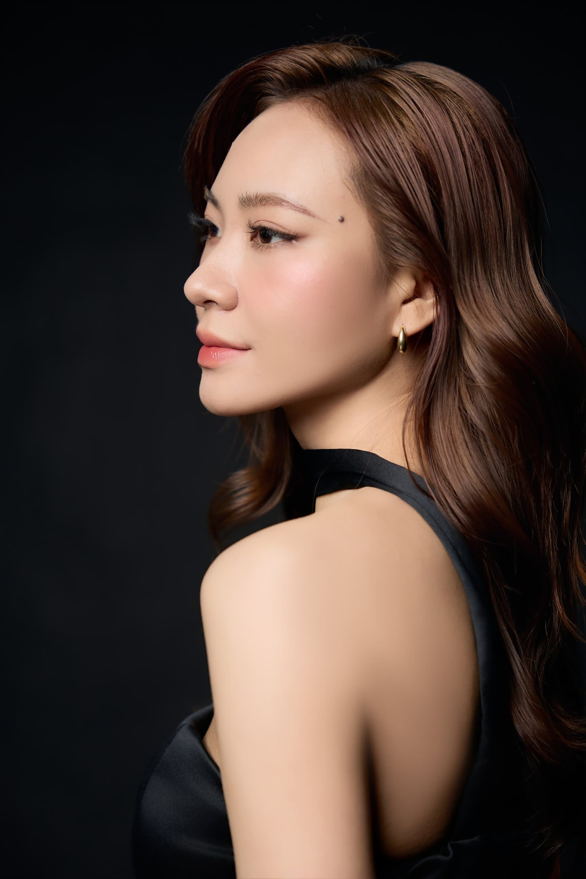 Khánh Trang quay nghiêng, vai trần, choker đen mảnh, gương mặt nhẹ nhõm trên nền tối — một người ít nói quen đứng ở phần quan sát