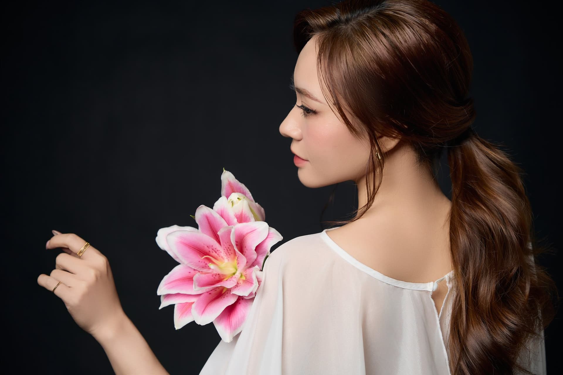 Cô gái áo trắng nhẹ, tay chạm vào bông ly hồng đang nở, gương mặt nghiêng nhìn xa — một lần đầu tiên, một kỷ niệm không cần nhân dịp