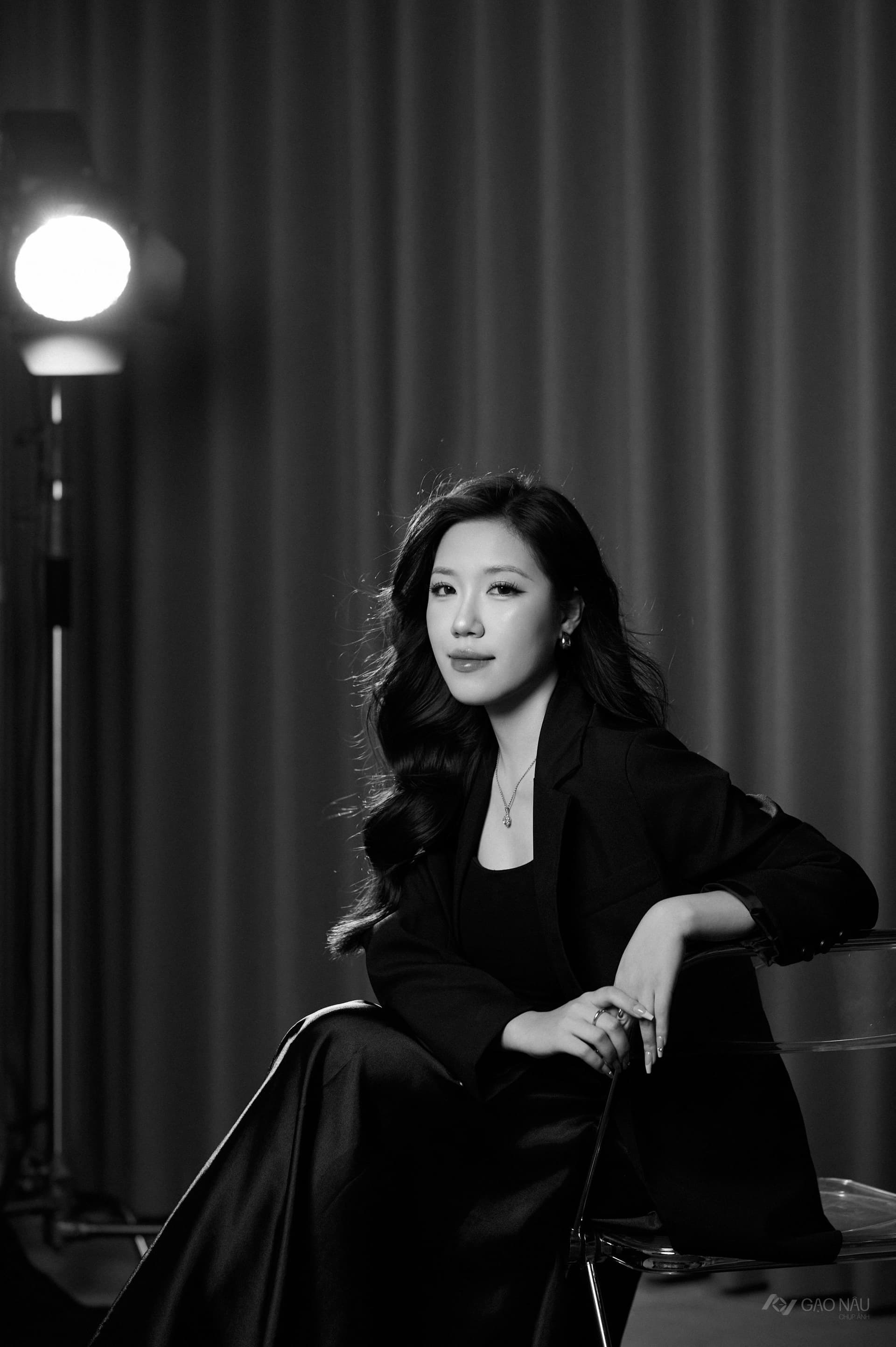 Người phụ nữ áo blazer đen, váy lụa đen, ngồi nghiêng trên ghế acrylic trong suốt, đèn spotlight studio đổ xuống vai, tone đen trắng