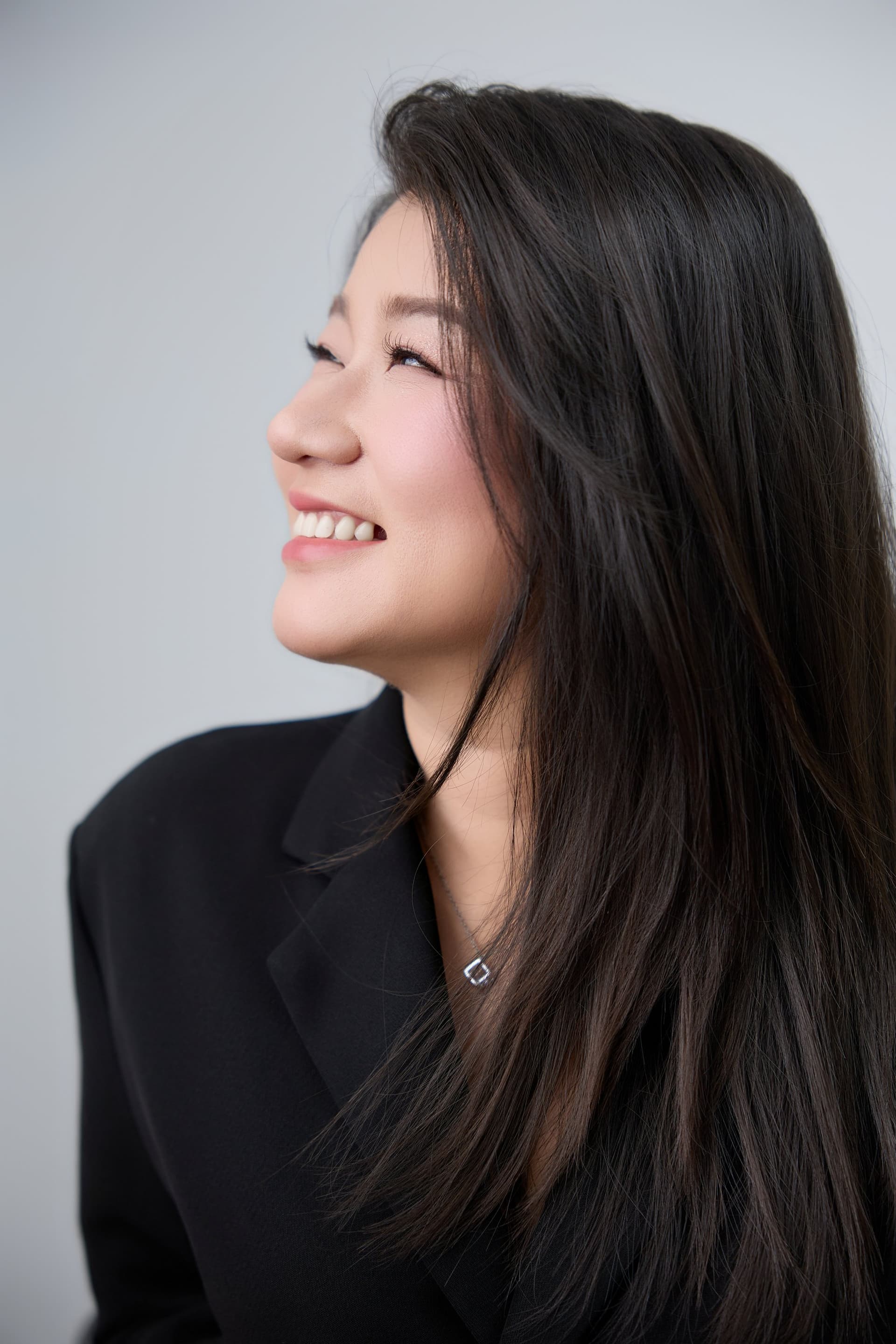Người phụ nữ ngồi tựa ghế, áo blazer đen, gương mặt suy tư — vai mẹ vẫn phải giữ sau khi chồng đi xa