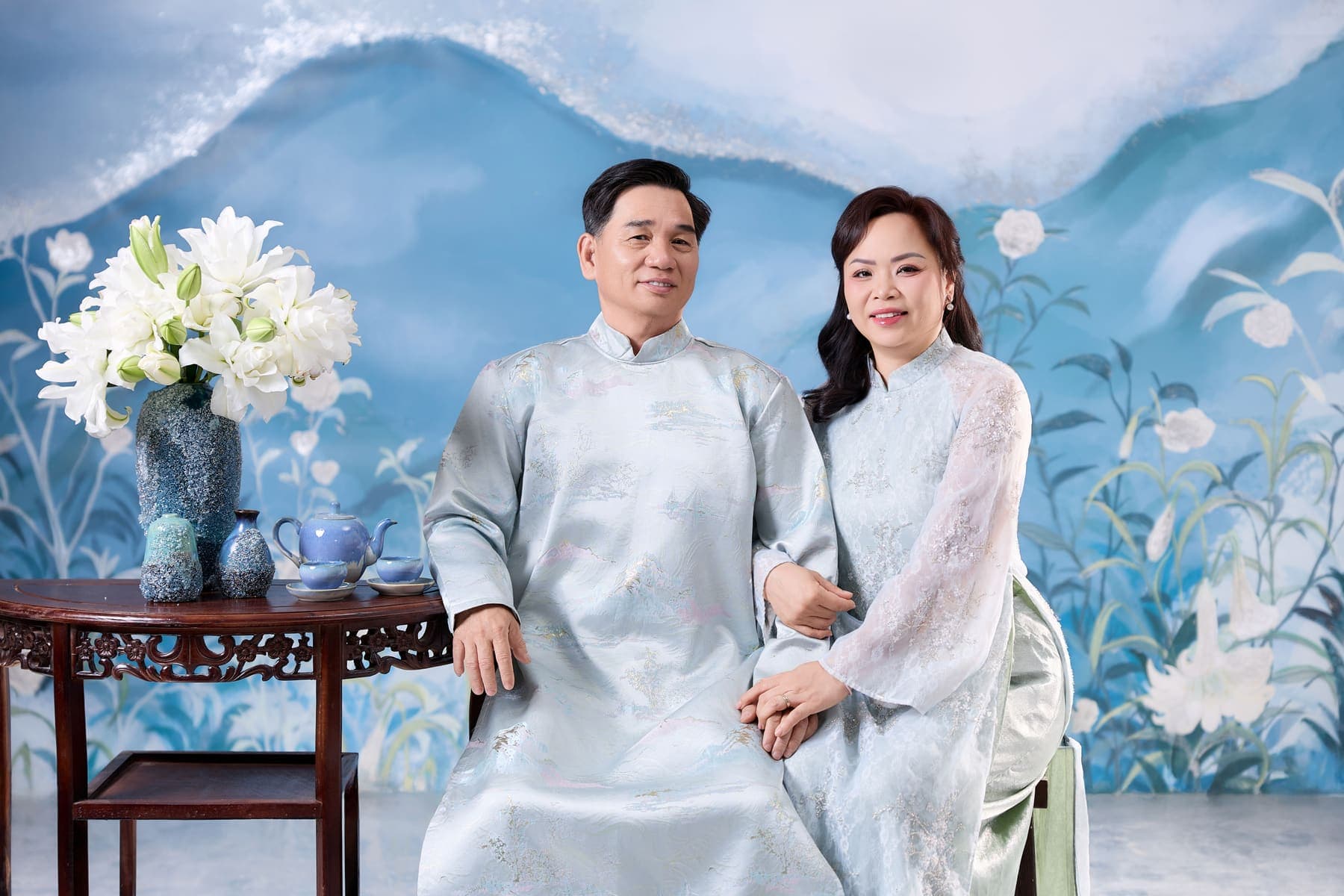 Hai vợ chồng trong áo dài xanh ngồi bên bàn trà, bình hoa trắng và nền núi lam dịu