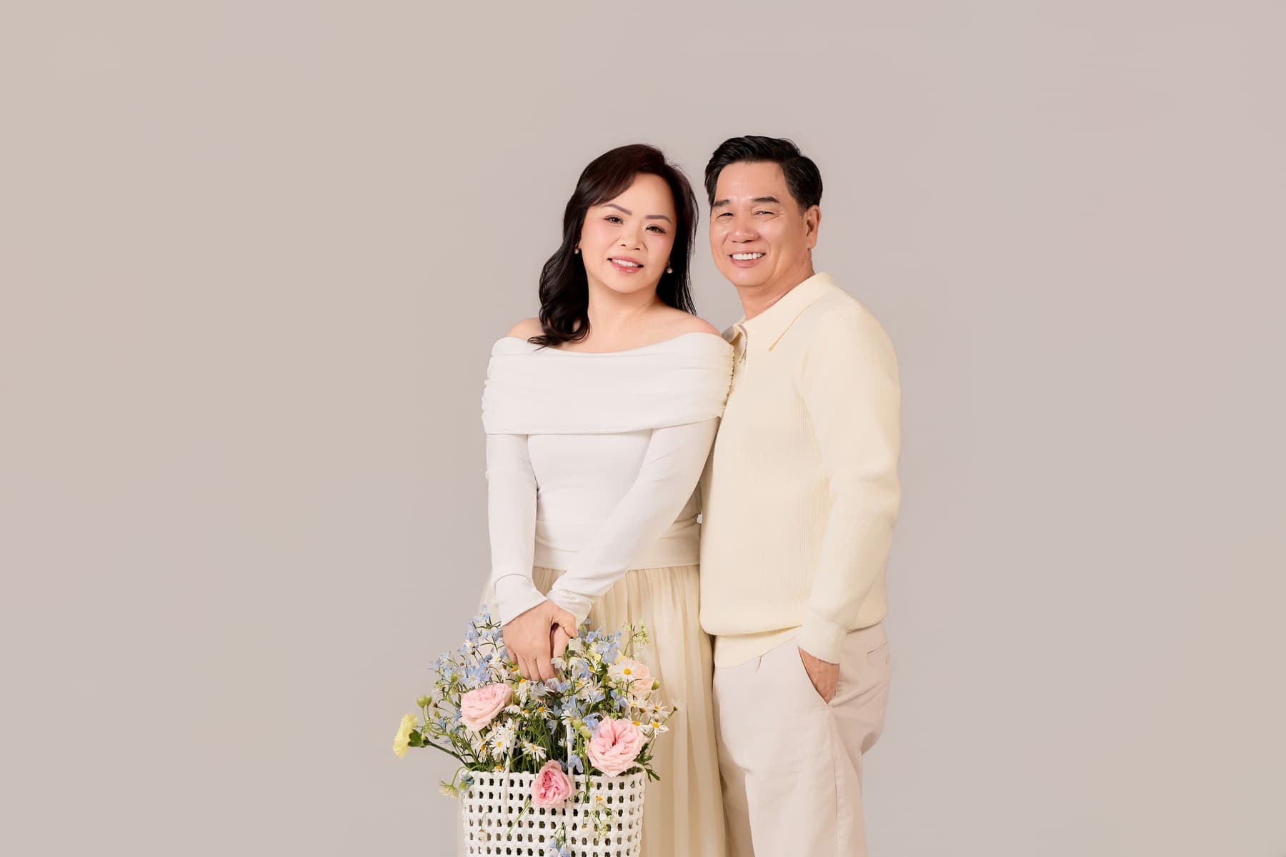 Hai vợ chồng đứng cạnh nhau trong trang phục sáng màu, bó hoa đặt phía trước