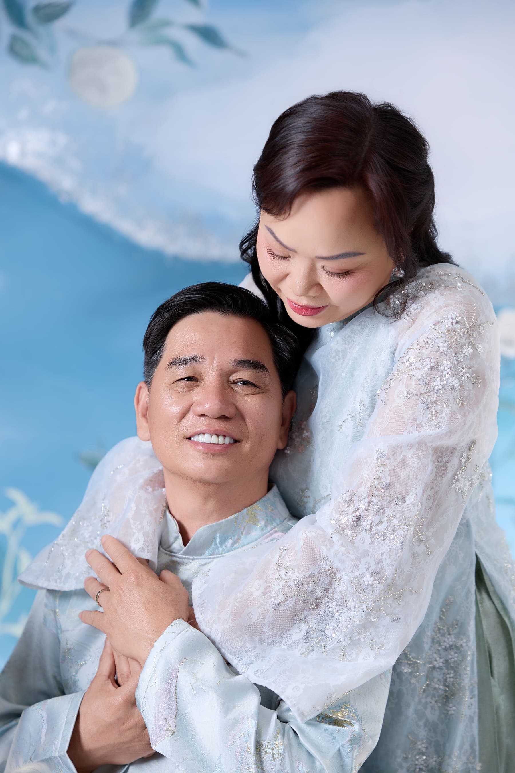 Người vợ ôm vai người chồng trong áo dài xanh, nụ cười hiền giữa nền hoa lam