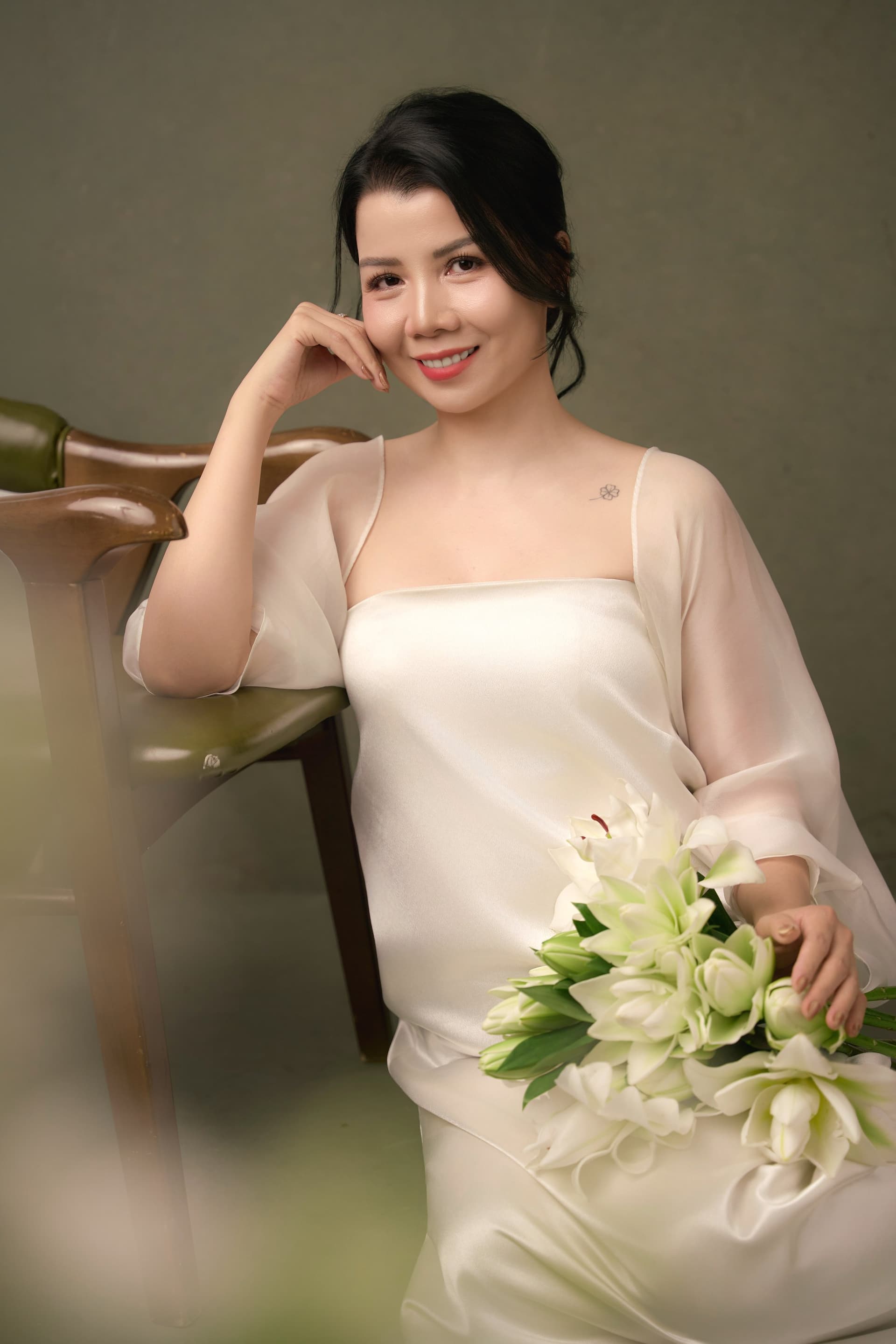 Chị Thanh Nhài ngồi trên ghế tựa, tay ôm bó hoa ly trắng rạng rỡ và mỉm cười nhẹ nhàng