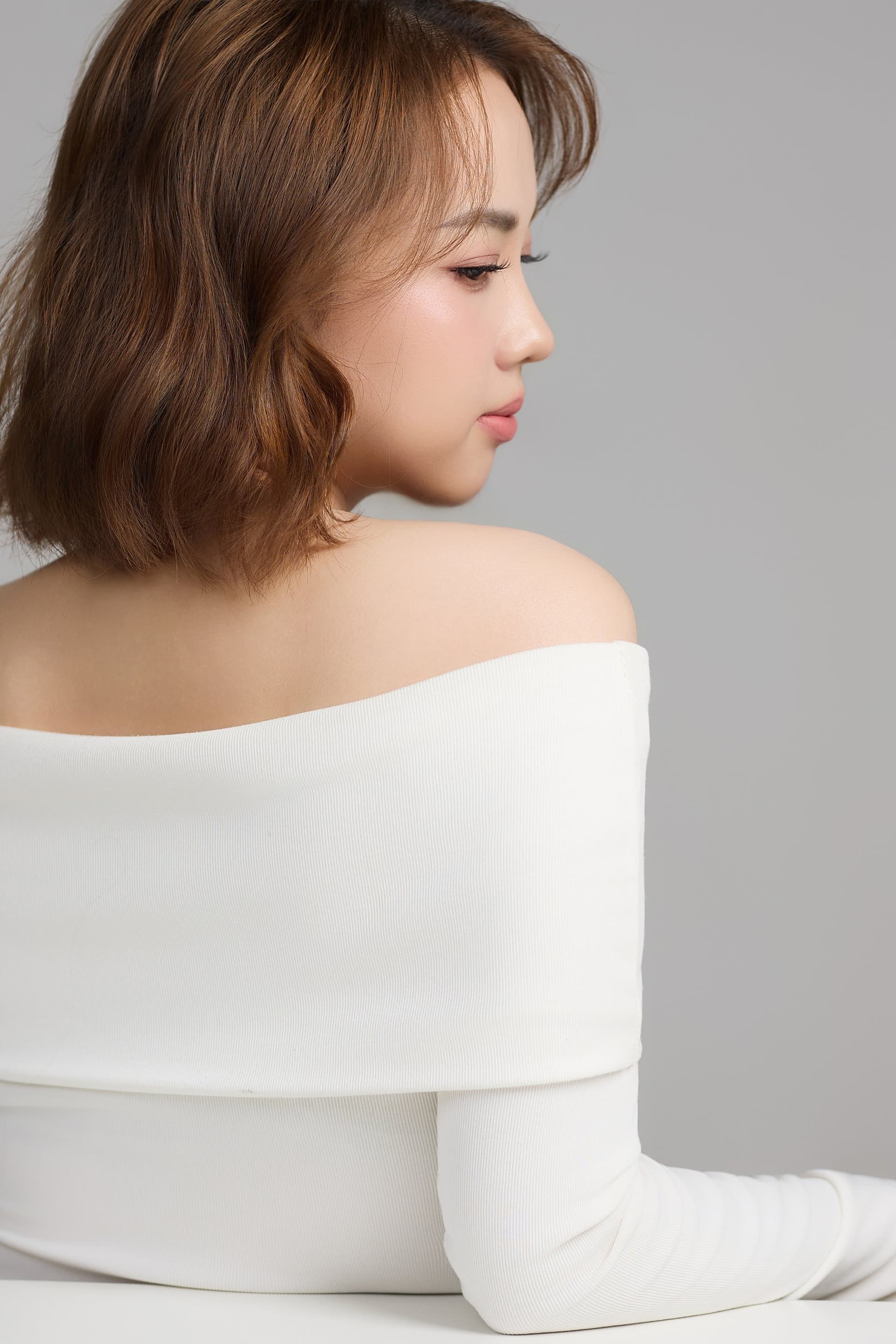 Người phụ nữ mặc áo trắng trễ vai nghiêng mình nhìn tránh chiếc gương, mang nét ưu tư đầy chiều sâu tâm trạng trước tuổi ba mươi