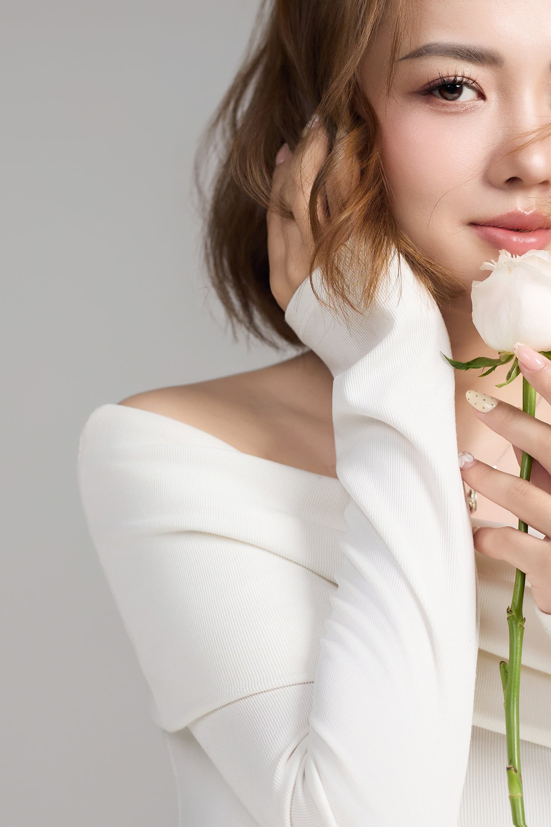 Cẩm Tú trong tà áo trắng trễ vai, hai tay nâng nhẹ nhành hoa hồng trắng sát ngực đầy dịu dàng
