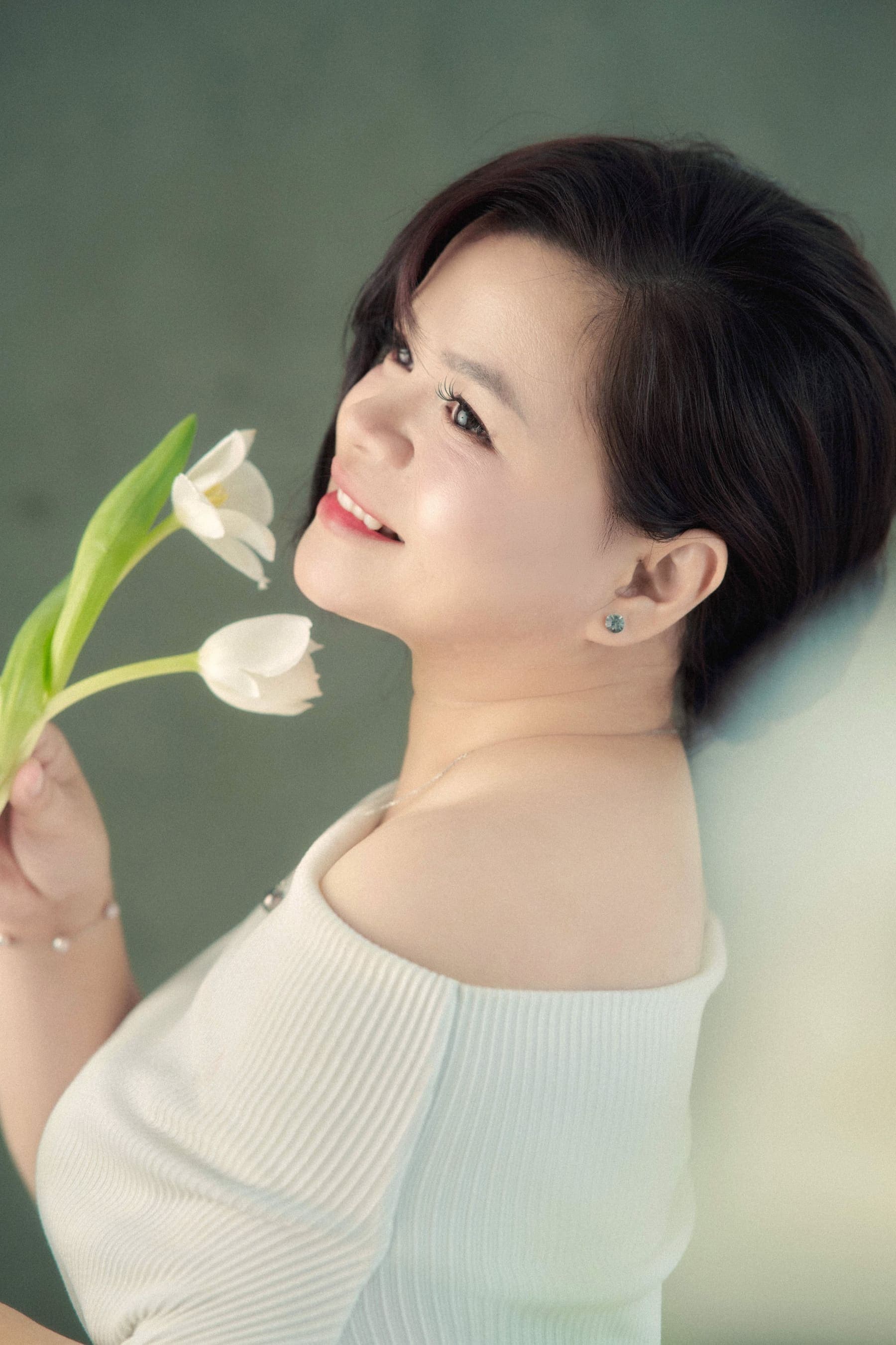 Người phụ nữ áo lễ trắng vai trần nghiêng đầu, mắt khẽ nhắm, ngửi bông tulip trắng — phần mềm vẫn còn đó, chỉ là đã quen được cất kỹ
