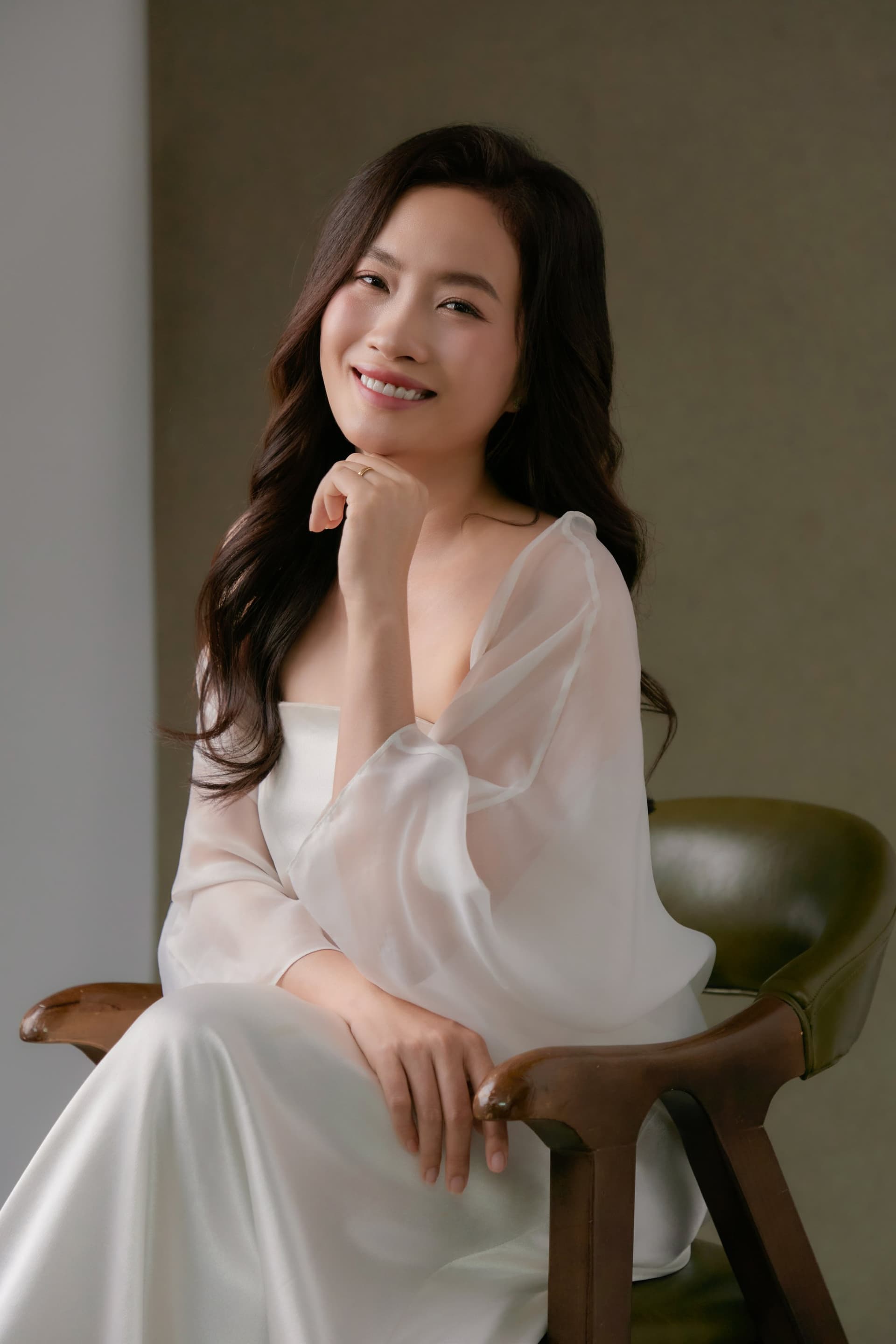 Lan Hương ngồi ghế gỗ tay tựa cằm, đôi mắt cười duyên dáng trong áo trắng nền