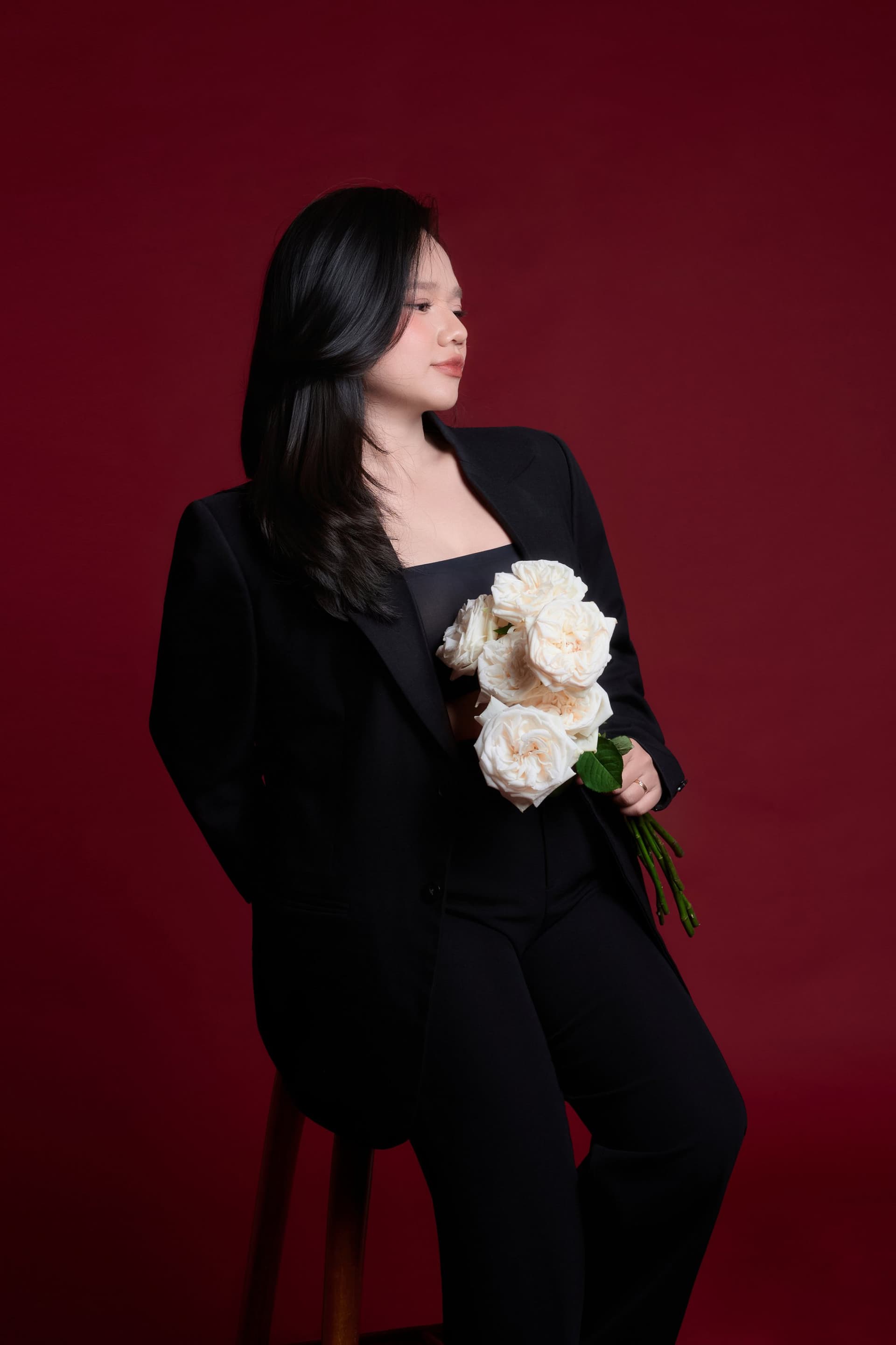 Ms Tiny Nguyễn thanh lịch trong bộ suit đen ngồi trên chiếc ghế đỏ, ánh mắt suy tư sâu lắng
