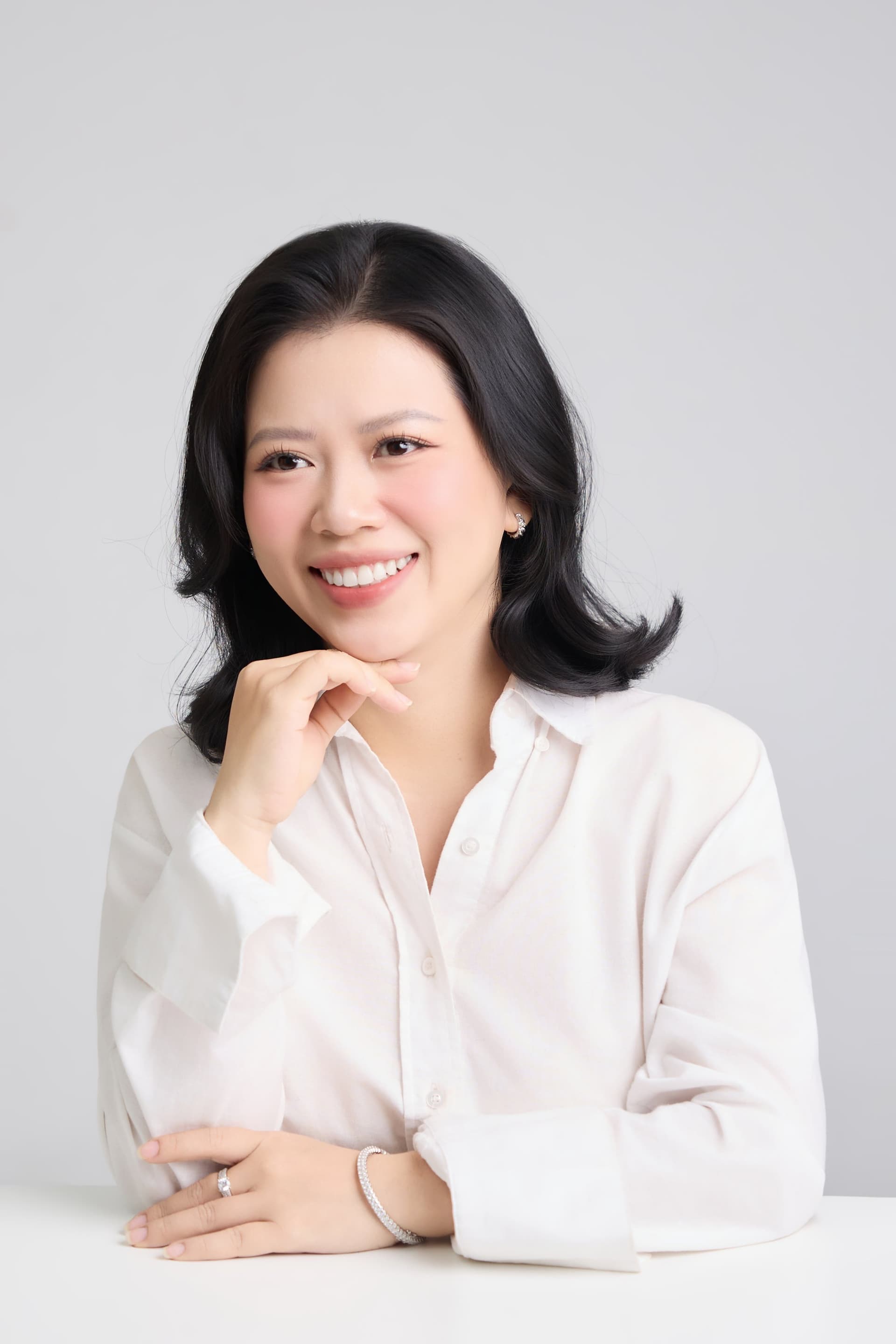 Người phụ nữ mặc sơ mi trắng mỉm cười nhẹ nhàng tựa tay vào cằm giữa không gian tối giản trầm ấm